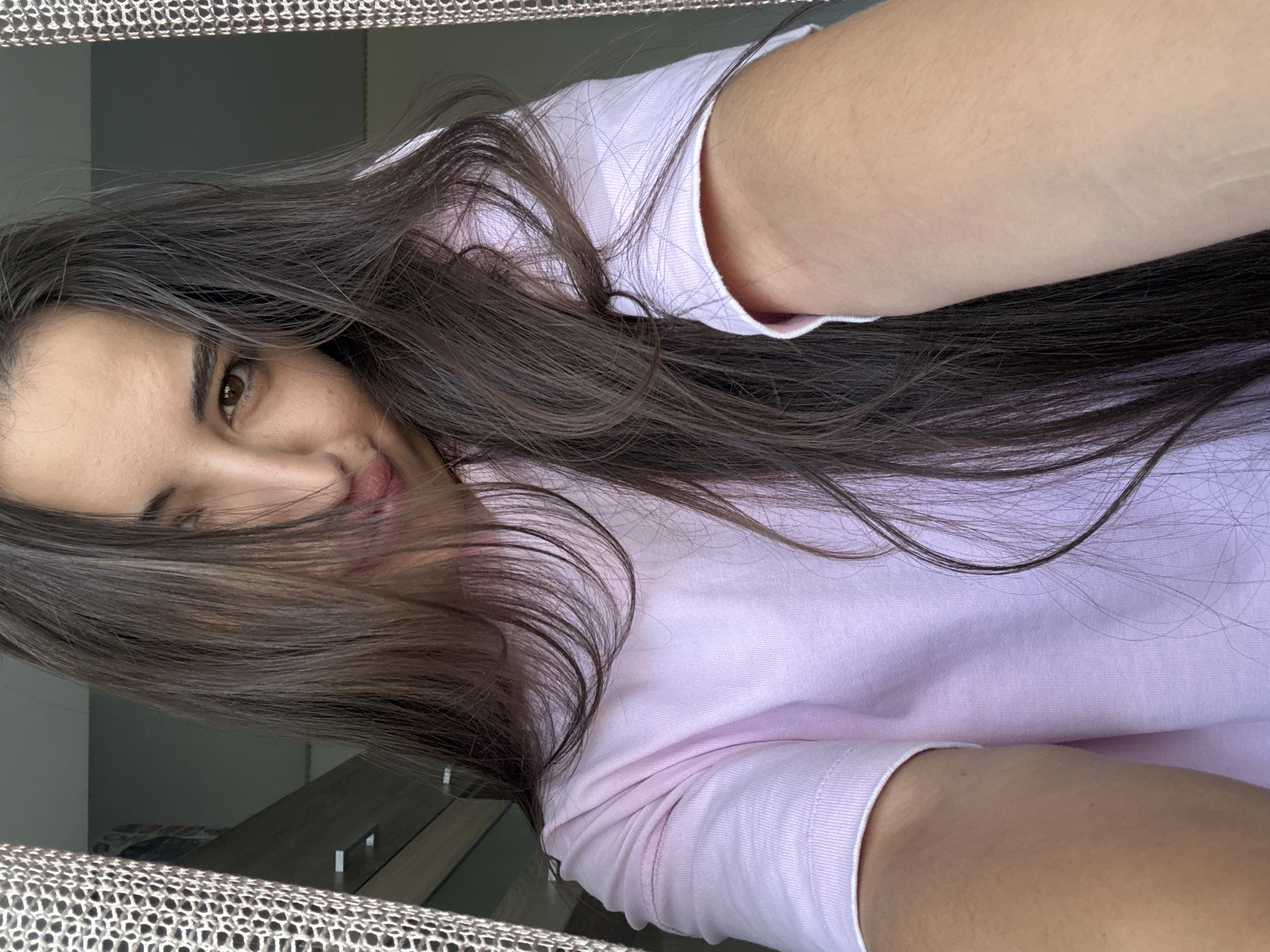 A young woman with long, dark brown hair is taking a selfie indoors. She is wearing a light pink t-shirt. Her hair is partially obscuring her face, and she is looking at the camera with a playful expression, her tongue slightly out. Behind her, a curtain with a textured pattern is visible to the left and right of the frame. The background appears to be a room with some furniture, possibly a dresser, visible in the upper left corner. There is no indication of the specific location within Chișinău, Moldova, or any environmental details that would pinpoint it to that city. The image captures a casual, personal moment.