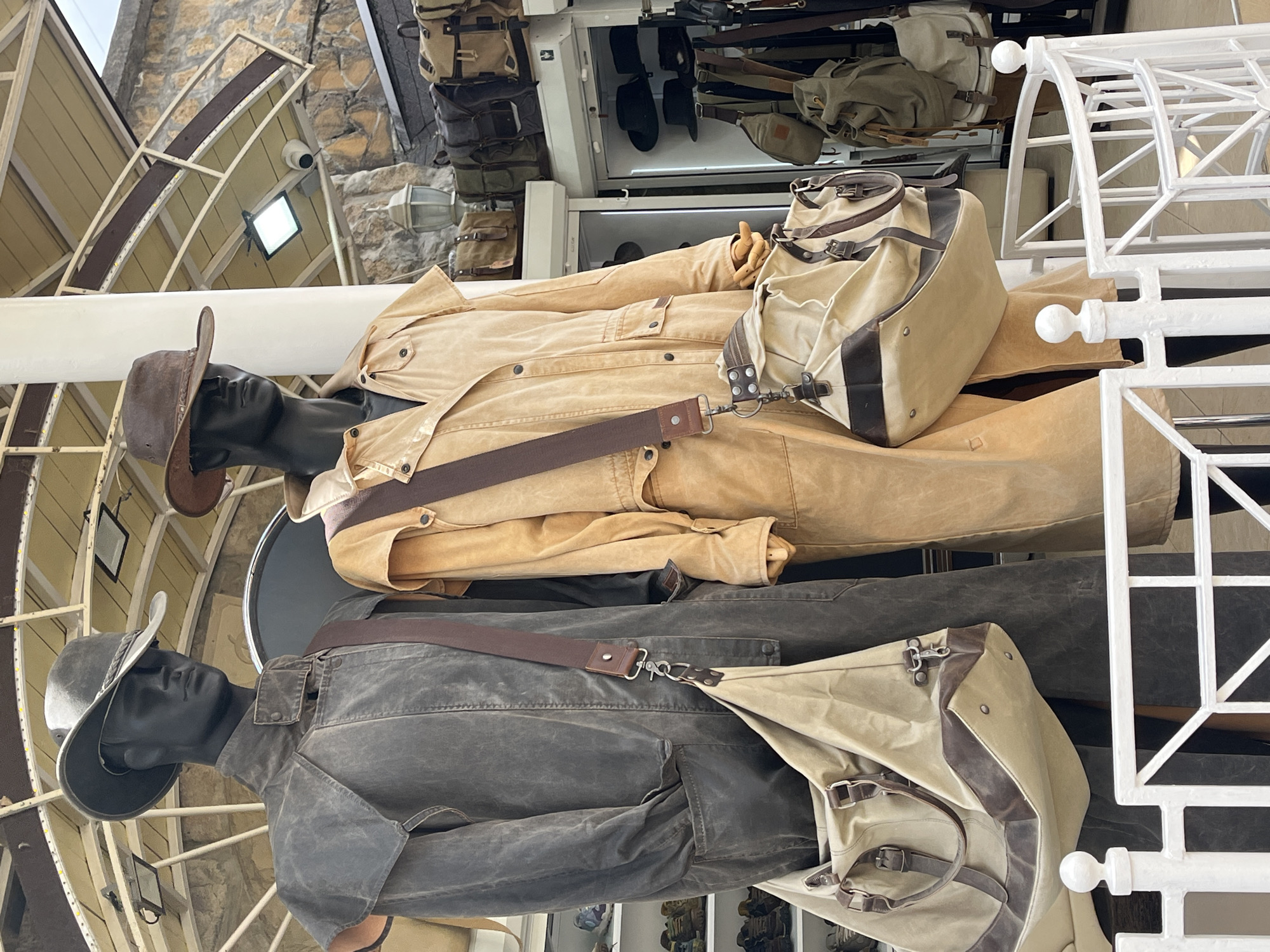 The image presents two mannequins dressed in outdoor attire, reminiscent of an explorer or adventurer, likely displayed in a retail setting in Kislovodsk, Russia. The top mannequin wears a light tan jacket and a brown cowboy hat, with a khaki duffel bag slung across its body. The lower mannequin sports a darker, gray-brown jacket and a gray wide-brimmed hat, also carrying a similar style khaki duffel bag. Both mannequins have dark skin and appear to be in profile.
The mannequins are positioned against a backdrop that includes architectural elements – a white decorative structure made of interconnected frames and balls - and shop fixtures, such as shelving displaying other bags and accessories. The location appears to be indoors, with an angled ceiling and visible stonework above, suggesting the character of the city.