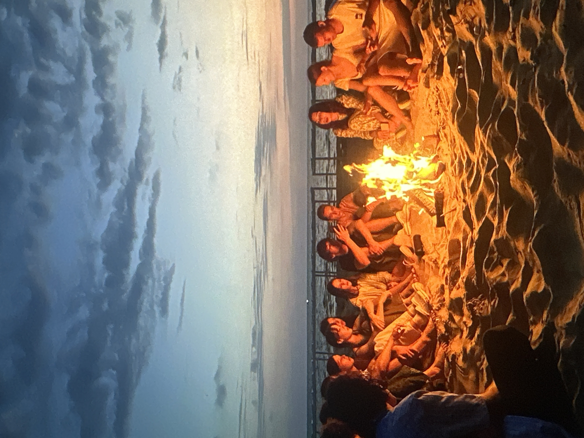 A group of young adults are sitting around a bonfire on a sandy beach at dusk. The sky above is a gradient of deep blue and orange, with scattered clouds. The ocean is visible in the background, with a faint railing or structure along the shore. The bonfire is bright and casts a warm, orange glow on the faces and clothing of the people surrounding it. Most of the individuals are dressed casually. Their postures suggest relaxation and conversation. The sand is textured with footprints and ripples from the wind. The overall scene depicts a social gathering in a natural outdoor setting during twilight. The location is not specified as being in Tallinn, Estonia.