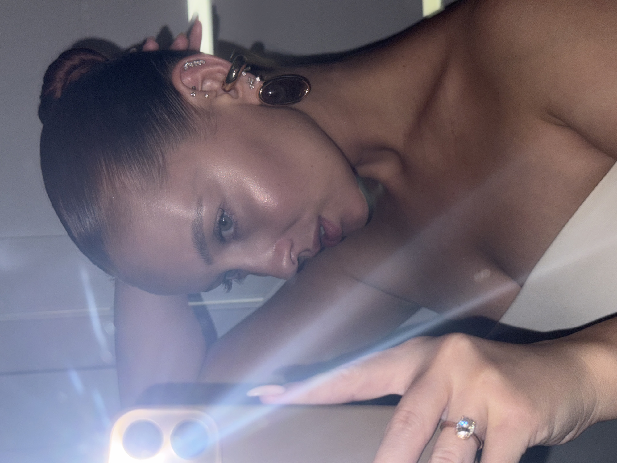 A woman is taking a selfie with a smartphone. She has dark hair pulled back in a bun, and multiple piercings in her left ear, including an oval-shaped earring. The phone's flash is illuminating the lower portion of her face and hand. A ring is visible on her left ring finger. The background is unclear. The location is not identifiable as the city of Chaikovsky, Russia.
