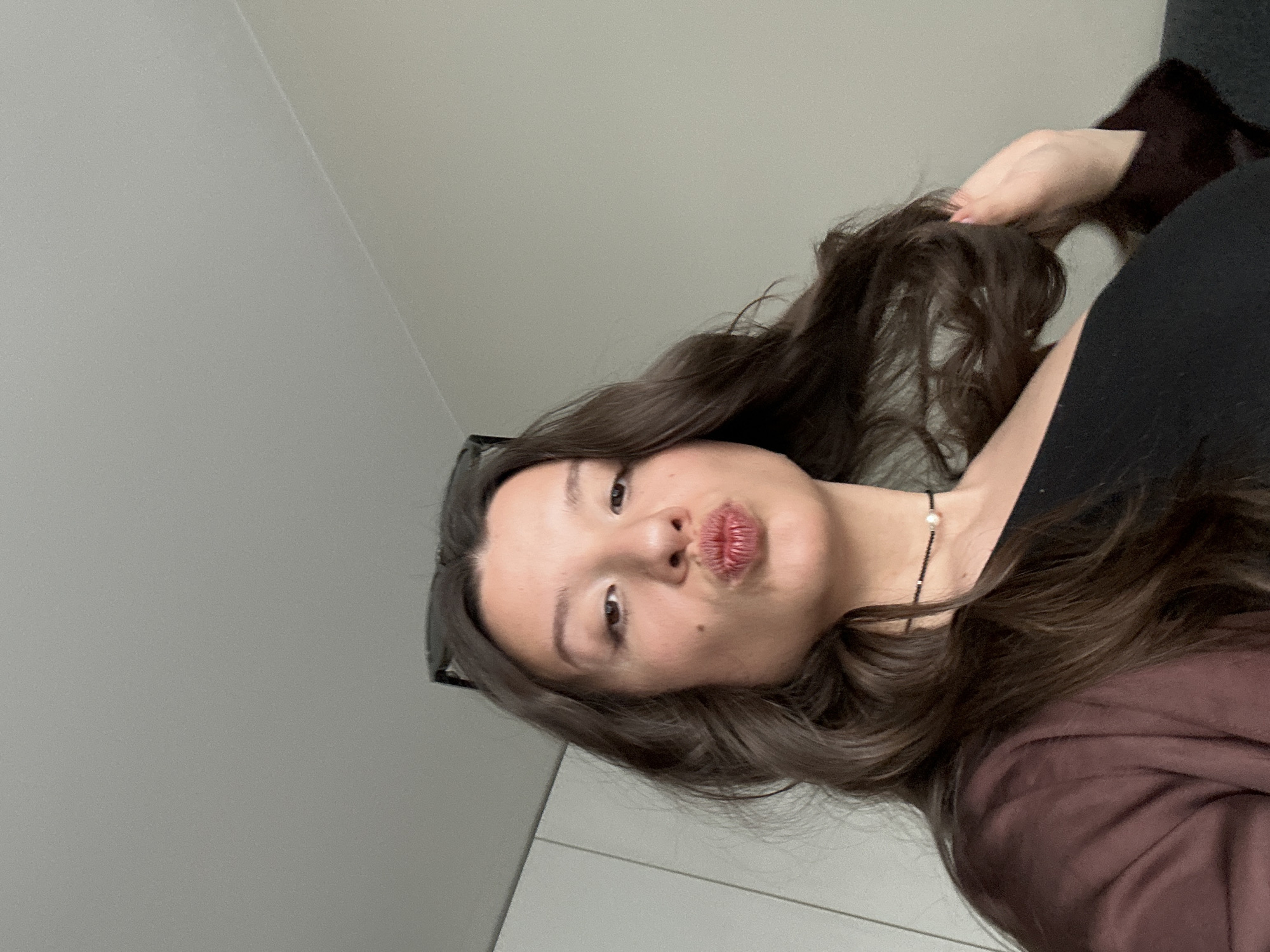 A young woman is posing for a selfie. She has long, dark hair and is wearing a dark-colored top. She is holding a strand of her hair in her hand and has a slight pout on her lips. A necklace is visible around her neck. The background is a plain white wall. There is no information available to confirm the location in Sofia, Bulgaria.