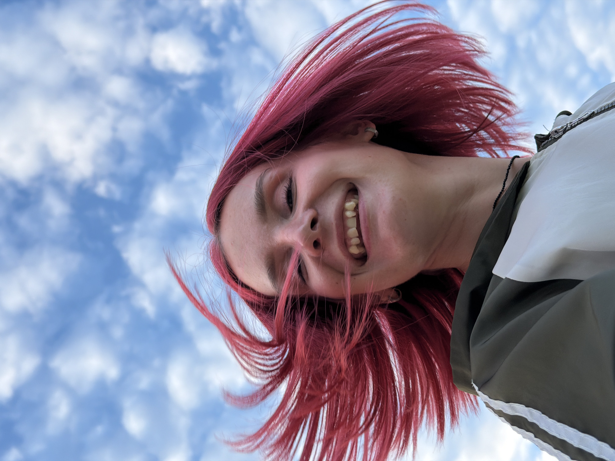 In an outdoor, low-angle shot, a young person with vibrant pink hair is captured mid-laugh against a backdrop of a bright blue sky dotted with white clouds. Their head is tilted back, and their hair is blown to the left by the wind, creating a dynamic effect. The person is wearing a dark green and white jacket with white stripes on the sleeves, and a black beaded necklace. They have a silver hoop earring in their visible ear. The overall impression is one of joy and freedom. The location is identified as Chaikovsky, Russia.