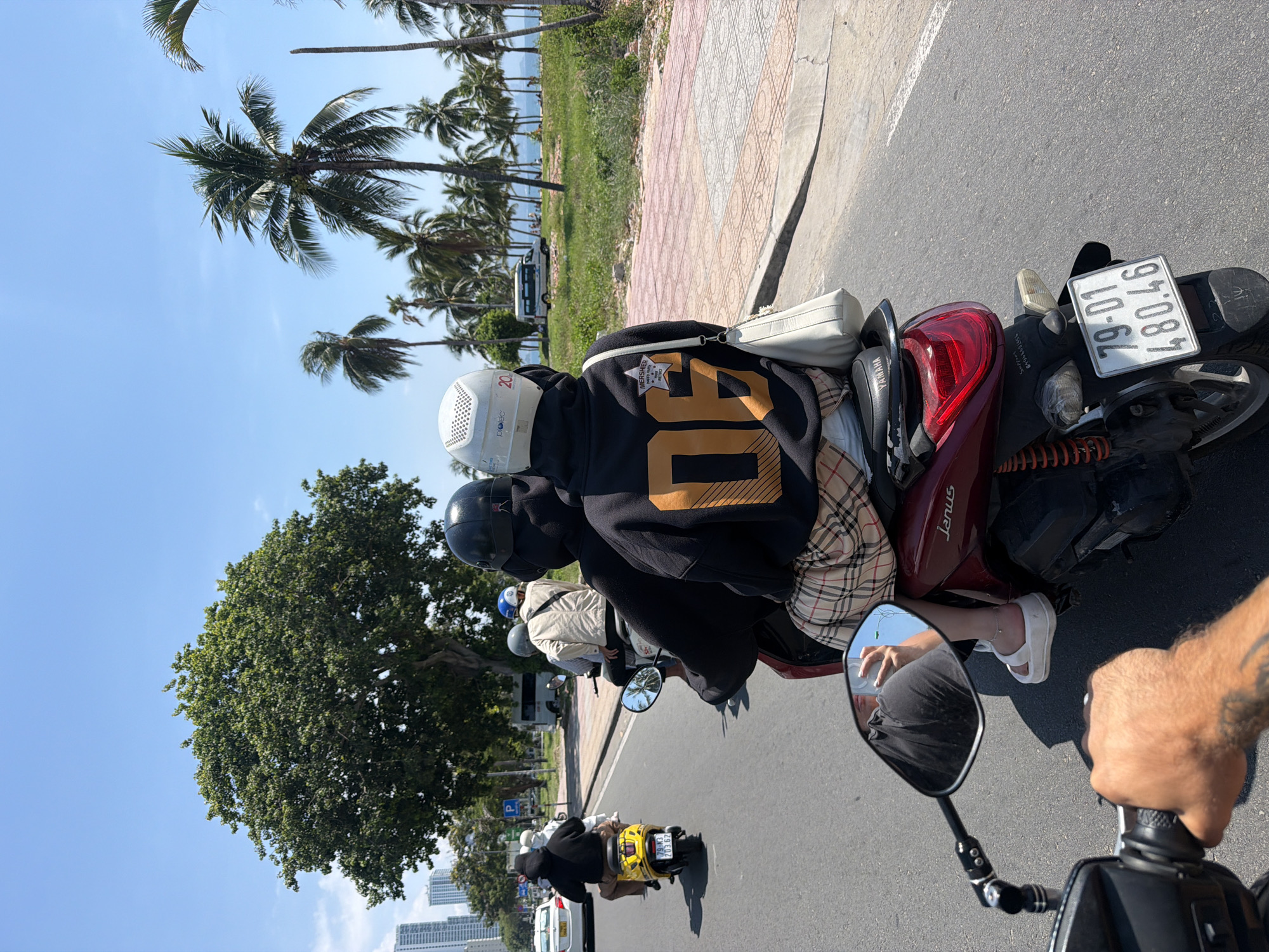 A group of people on scooters ride down a street in Nha Trang Ward, Vietnam, on a sunny day. The foreground shows a hand on a scooter's handlebar, with a tattooed forearm visible. Behind this, a person wearing a black hoodie with orange "06" lettering and a plaid skirt is seated on a red scooter. Another scooter with two people, one in a white helmet, is ahead. Further down the road, a yellow scooter with a rider is visible. Palm trees and a large tree line the street, with buildings and the ocean in the distance. A white van is parked on the side of the road. The license plate on the red scooter reads "79-D1 480.46".