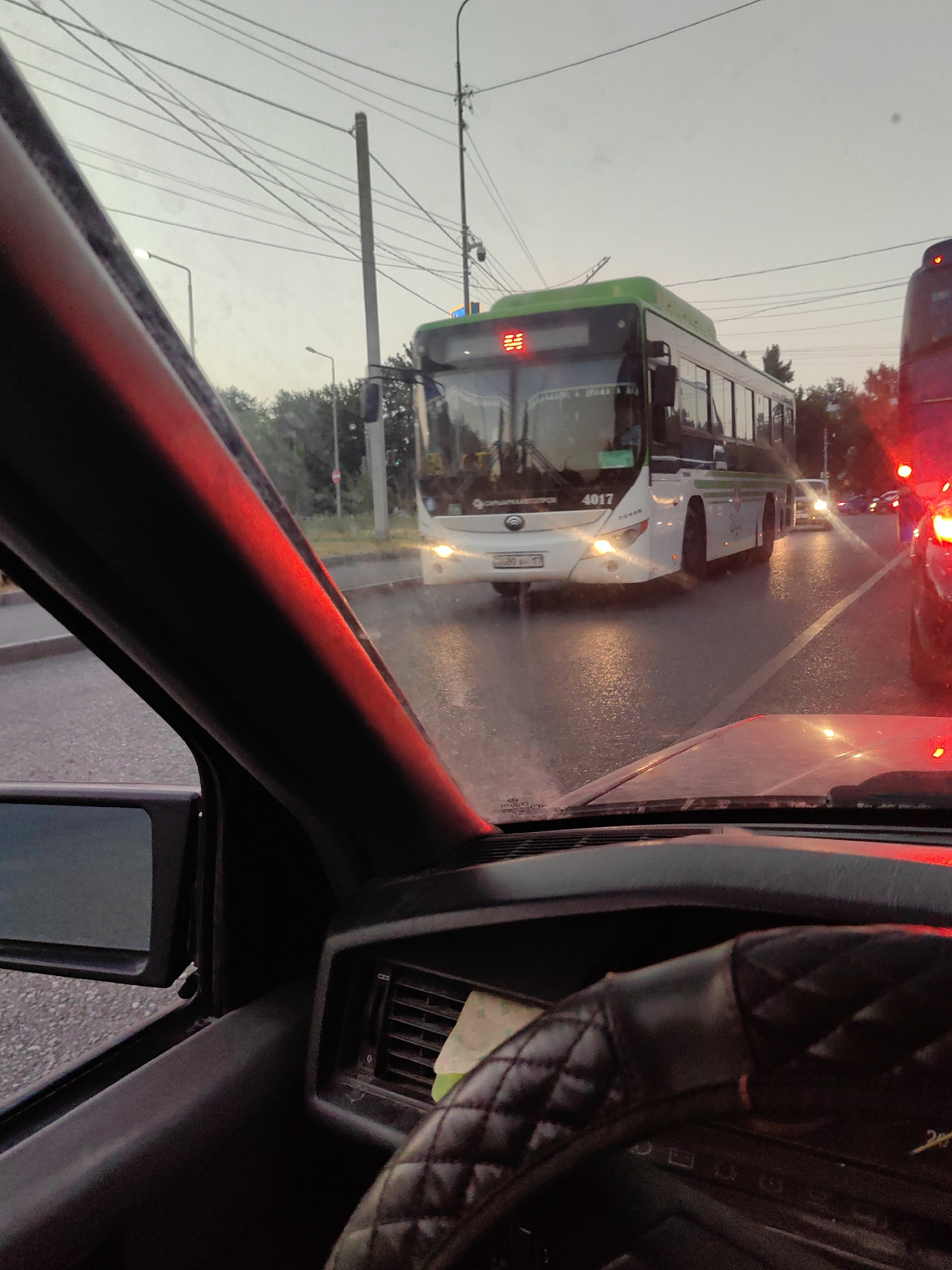 The image captures a view from inside a vehicle, looking forward through the windshield. In the foreground, a portion of the car's dashboard and steering wheel are visible. The steering wheel is covered in a diamond-stitched black leather material. To the left, the driver's side mirror is also visible.
The main subject of the image is a green and white bus, identified by the number "4017" on its front, which is ahead of the vehicle from which the photo is taken. The bus has its headlights on, and the wet road surface reflects the vehicle's lights and the surrounding streetlights. Another vehicle, possibly a bus with red taillights, is visible to the right.
The environment appears to be an urban setting with streetlights and power lines overhead. Trees line the side of the road in the background. The sky is overcast, suggesting it might be dusk or a cloudy day. The presence of wet roads indicates recent rain or a damp environment. Based on the visible license plate format and the presence of such buses, the location is likely Shymkent, Kazakhstan. There are no people clearly visible within the frame. The scene depicts traffic on a city street.
