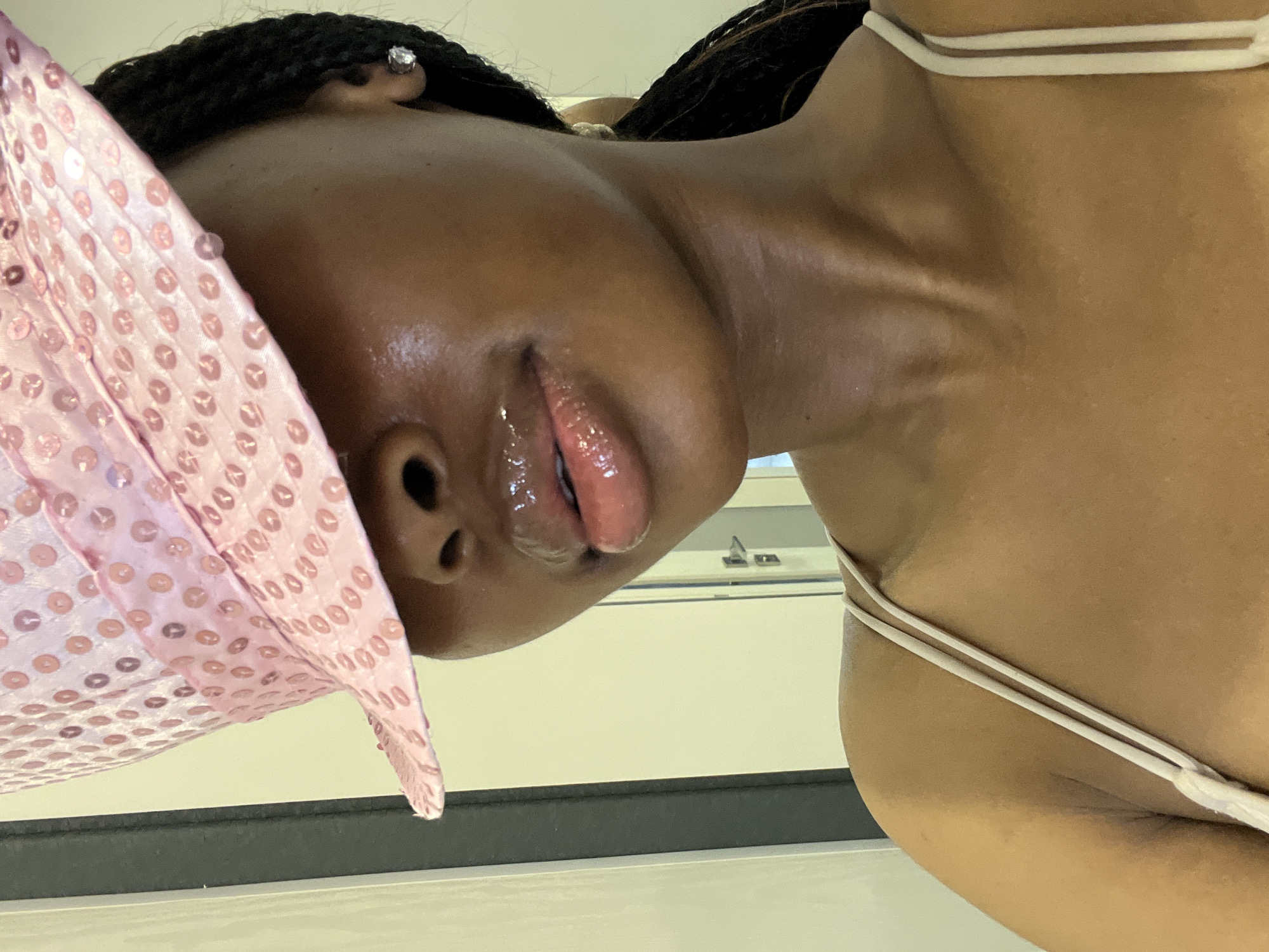 A close-up, slightly angled, medium shot shows the upper body and face of a young Black woman wearing a light pink sequined bucket hat that obscures her eyes. The hat has a band of lighter pink fabric. Her lips are glossy and slightly parted. She has on a light-colored tank top with thin straps. The background is out of focus, appearing to be an indoor setting with a wall and door visible. The lighting suggests a bright, indoor environment, possibly in Kazan, Russia, given the context.