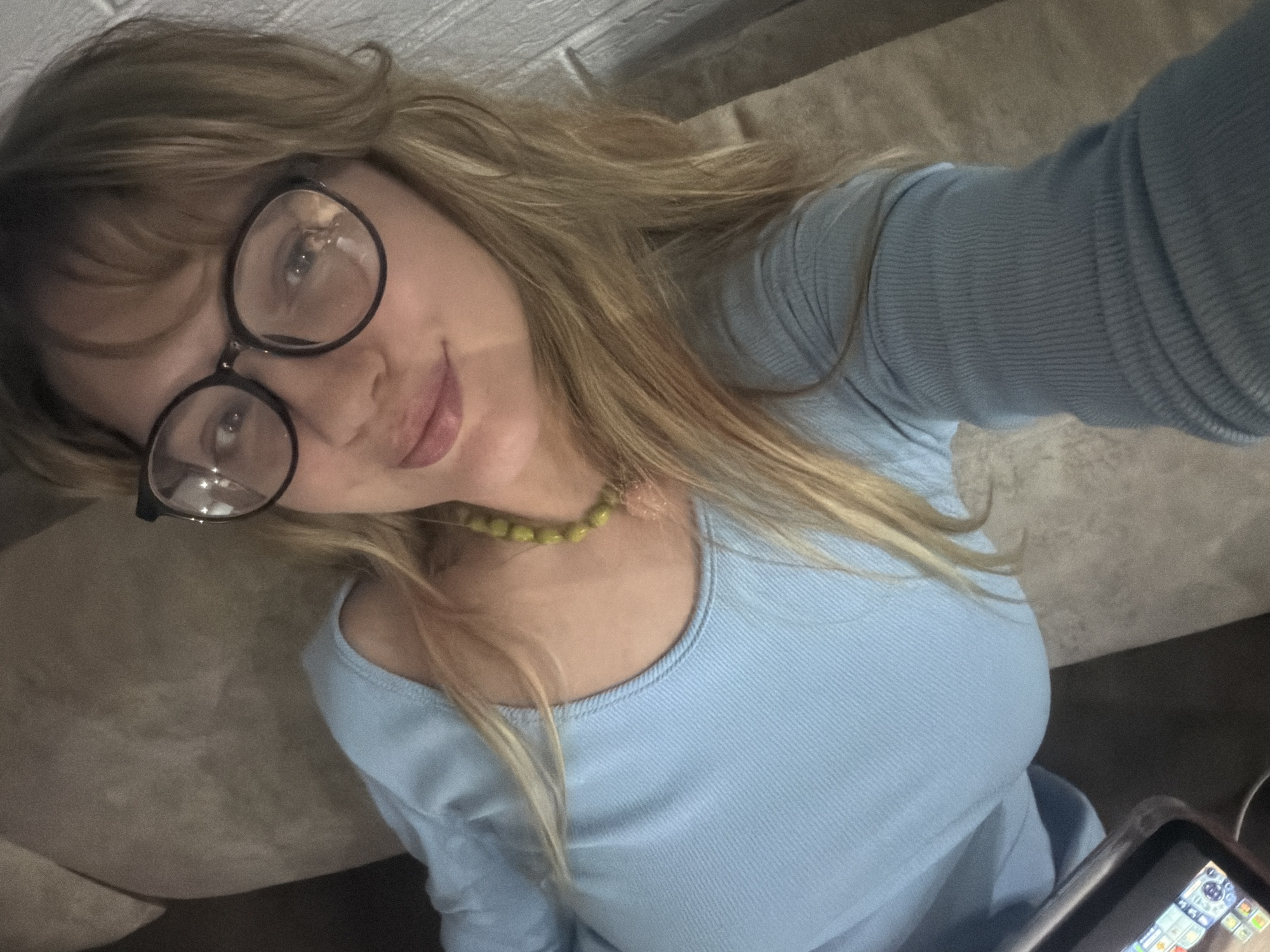 A woman with blonde hair and glasses is lying down in Большая Шильна, Russia. She is wearing a light blue long-sleeved shirt and a green beaded necklace. She is holding a cell phone. The background is blurry and indistinct.