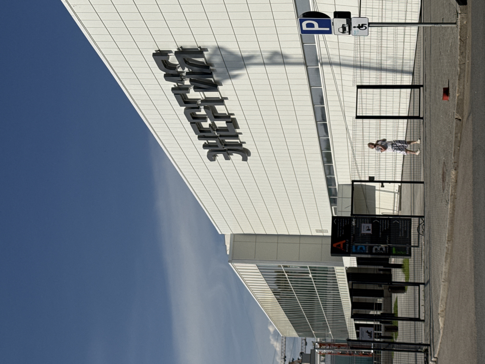 On a sunny day in Perm, Russia, a man walks on a sidewalk beside a modern building, identified by its signage as "Dymova." The building has a white tiled facade, reflecting the sunlight, with a clear blue sky visible above. A parking sign featuring a "P" and accessibility symbol is near the building's entrance. Various black signage structures stand along the sidewalk. In the background, glimpses of other urban structures and construction cranes can be seen.