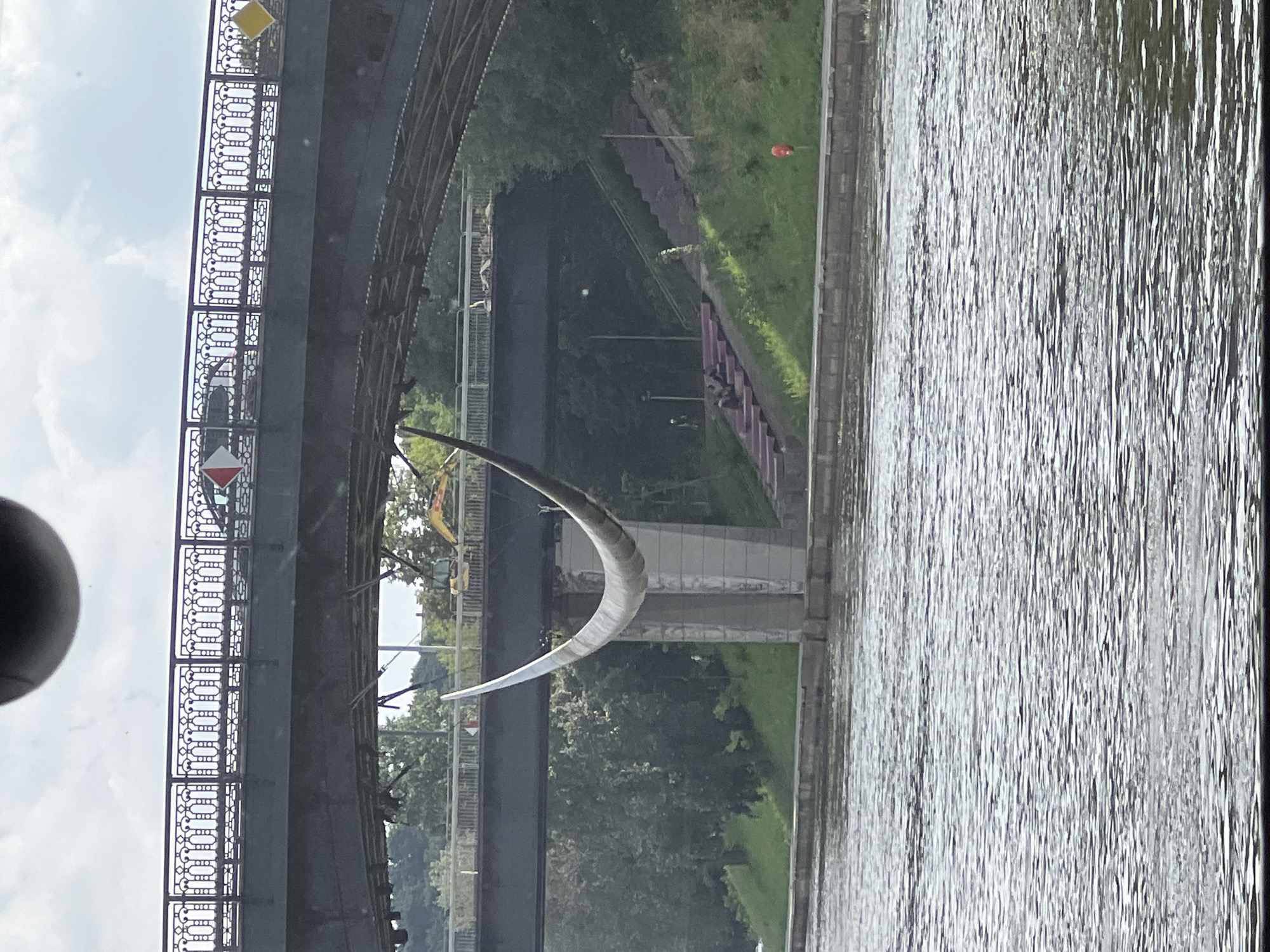 The image shows a large, curved metal sculpture suspended between two bridges over a body of water, likely the Neris River in Vilnius, Lithuania. The sculpture is pale in color and appears to be a thin, elongated arc. The upper bridge, a darker grey structure with ornate railings, has vehicles visible on it, suggesting it is a road bridge. Below this, a second, wider bridge is visible, with its underside showing construction or repairs. On the grassy embankment to the right of the lower bridge, a set of red and grey stairs leads up the slope, with several people seated on the stairs. A red cylindrical object, possibly a buoy or a marker, is visible near the base of the embankment. Trees and greenery surround the area, and the sky is partly cloudy. In the background, a yellow excavator is visible on the embankment, indicating possible ongoing construction or maintenance work. The water in the foreground is choppy and reflects the overcast sky.