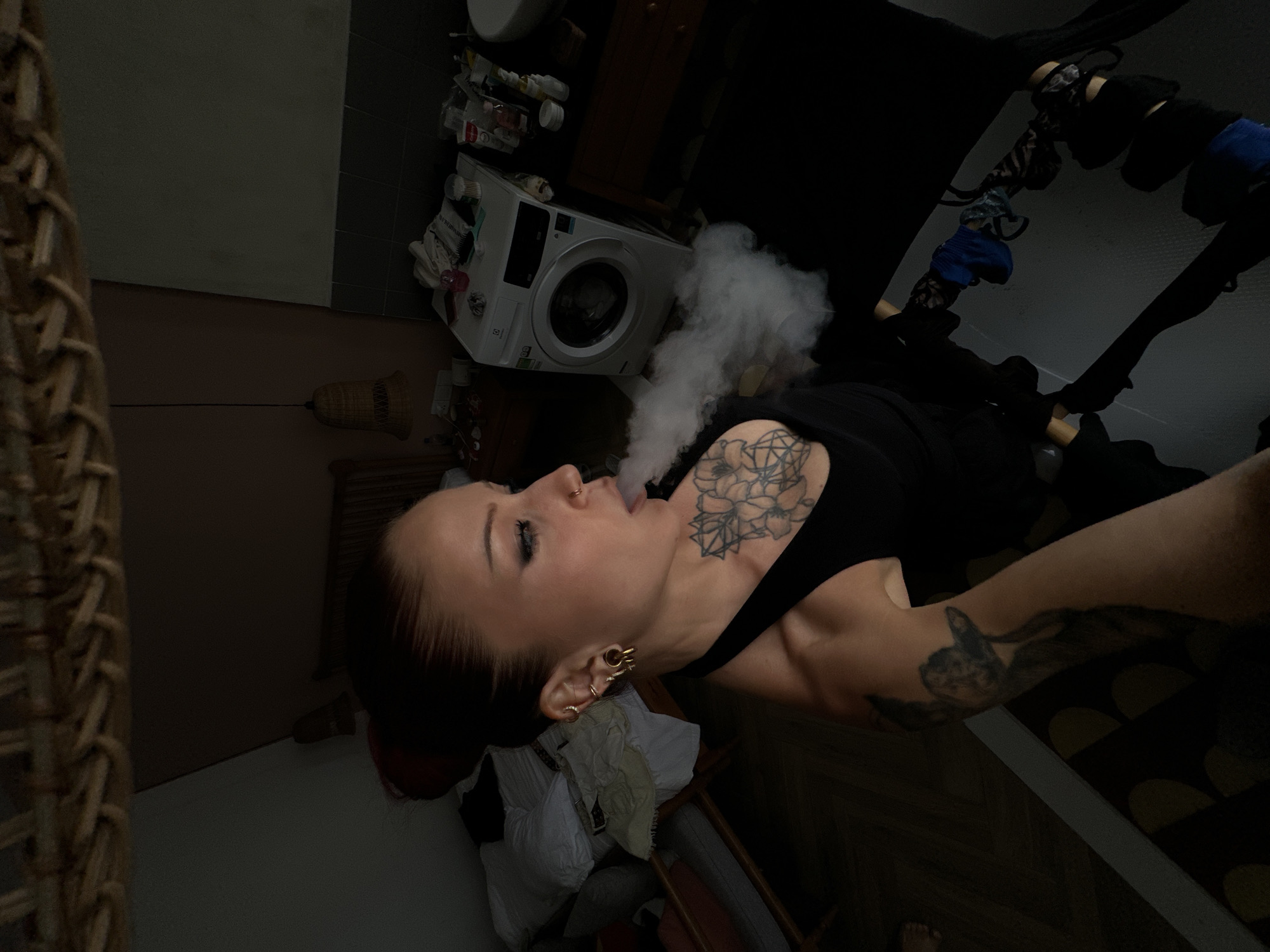 A woman with red hair in a bun, wearing a black tank top, exhales a cloud of vapor. She has extensive tattoos on her chest and left arm. Her ears are adorned with multiple piercings, including large gold plugs. She is facing to her left, with her mouth open, and appears to be in an indoor environment. In the background, a white washing machine is visible, along with various toiletries and household items on a shelf. A bed with white bedding is partially visible to the left. The lighting is dim, casting long shadows. The scene is not identifiable as being located in the city of Đà Nẵng, Vietnam.