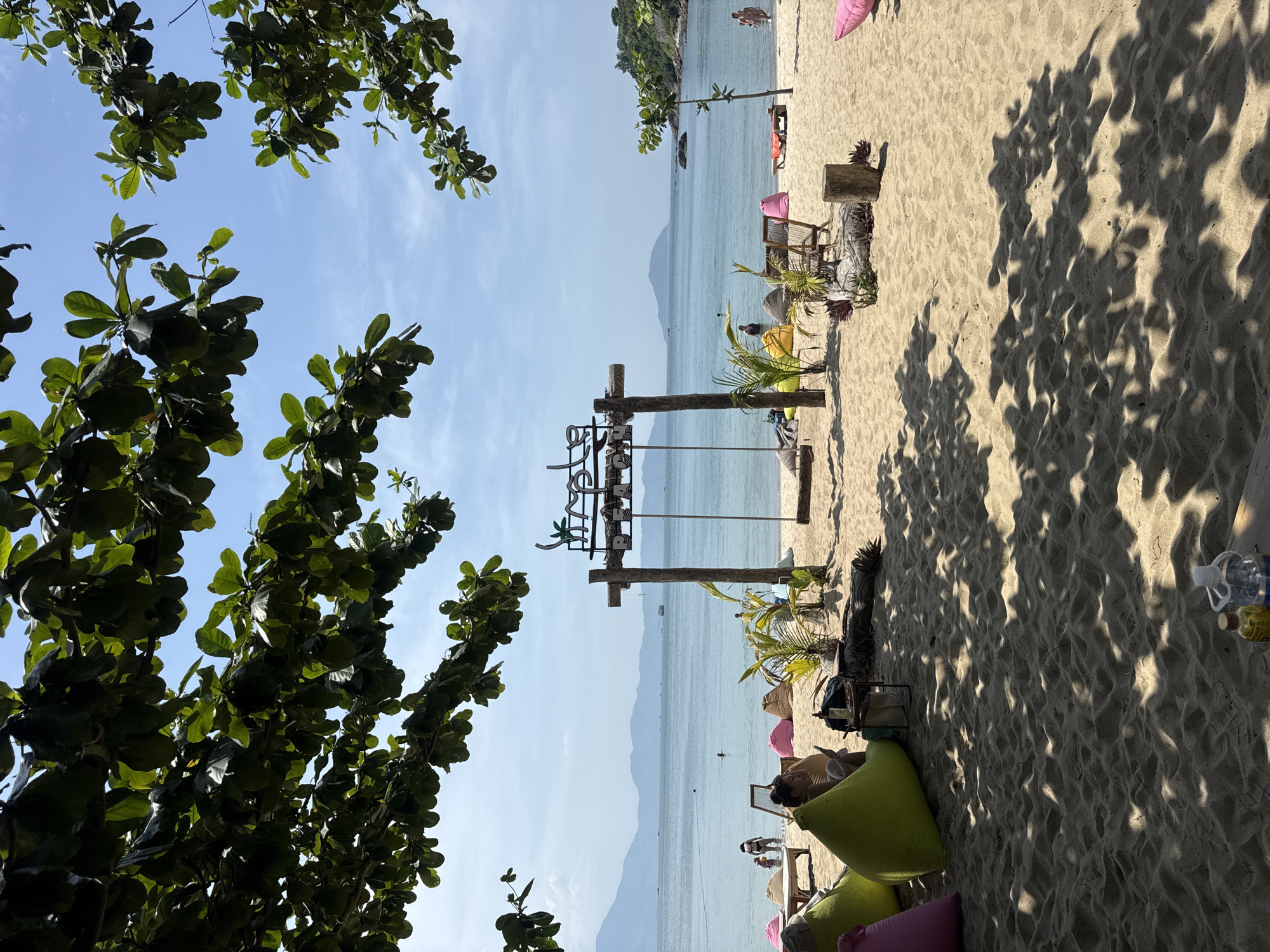 The image depicts a beach scene at an unknown location. The foreground features tree branches with green leaves on the left side. The beach itself is sandy and sparsely populated with beach chairs, beanbag chairs, and some wooden structures, including a sign. In the background, the ocean stretches out to meet the horizon, with distant landmasses or islands visible. A few people are present, some standing on the beach and others in the water. The sky is mostly clear and light blue. The lighting suggests it is daytime, with shadows cast across the sand.