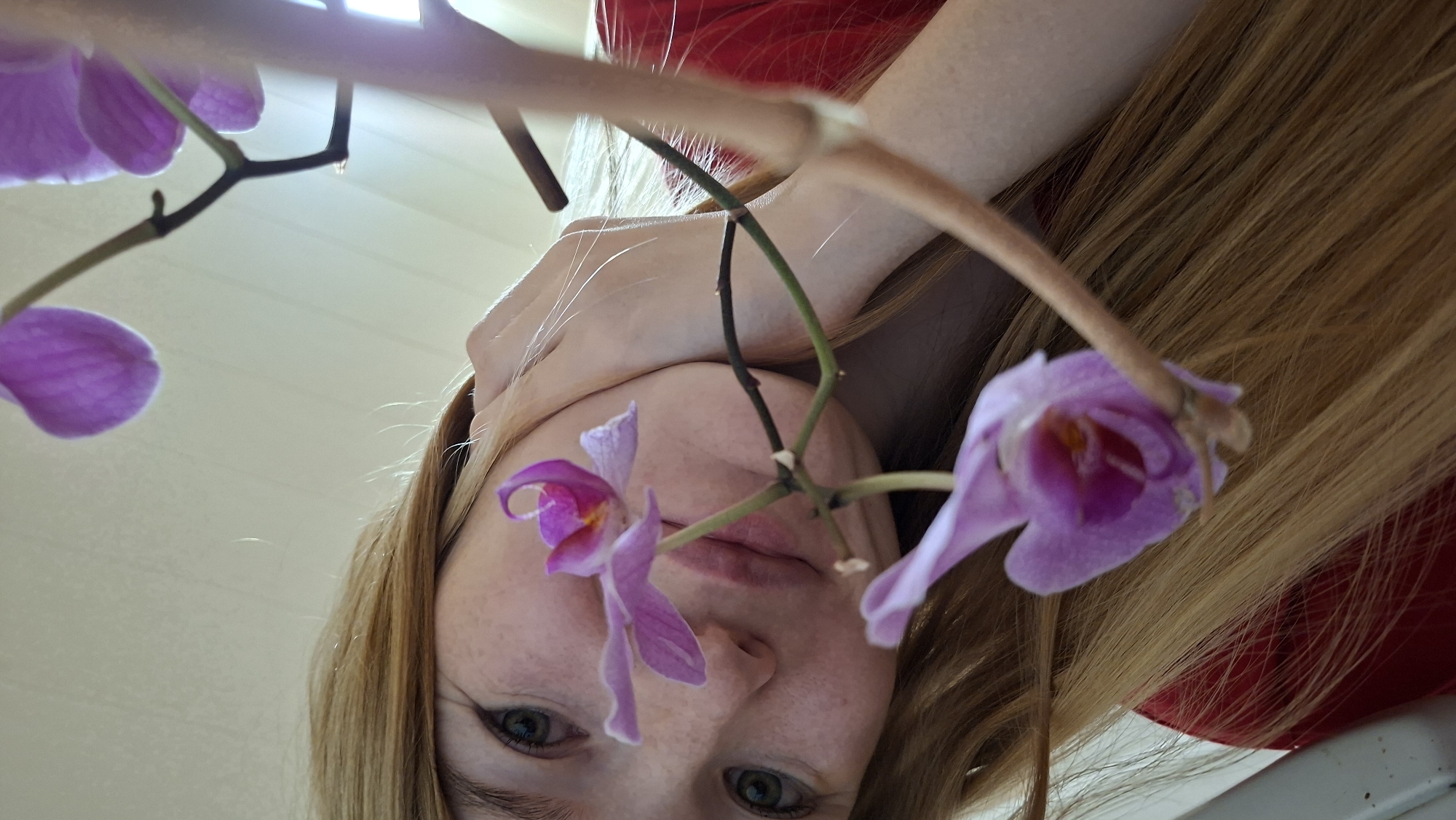 The image is a close-up, low-angle selfie of a young woman with fair skin and freckles, and long blonde hair. She is wearing a red shirt. She is holding a pink orchid with her right hand, which is resting on her neck. Her left eye is visible and appears green. The background is a plain, light-colored wall. The image appears to have been taken indoors. The location is specified as Bolshoy Bukor, Russia.