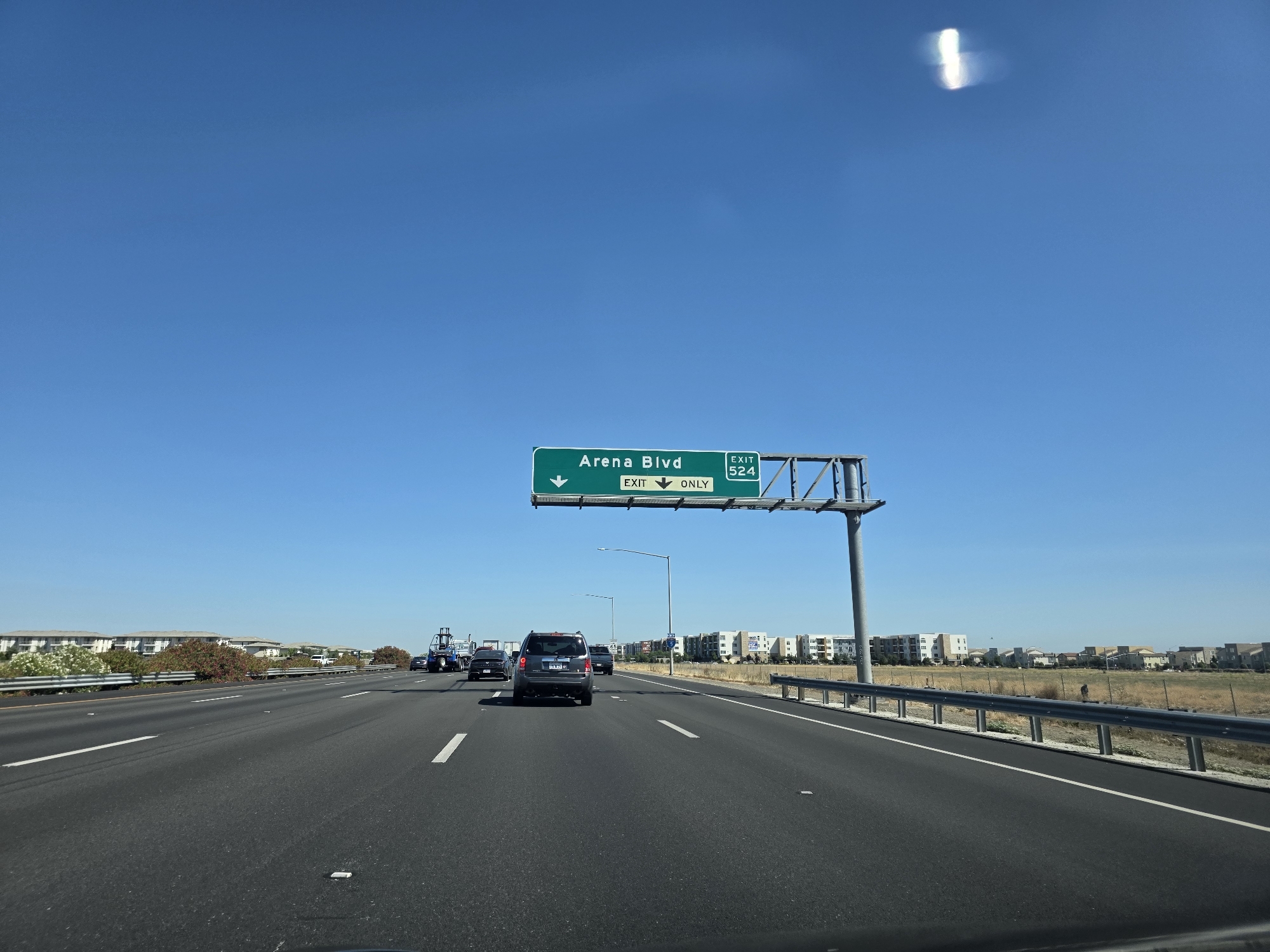A clear blue sky with a lens flare anomaly is visible above a multi-lane highway in Sacramento, California. A green overhead highway sign indicates "Arena Blvd" and "EXIT 524" with an arrow pointing down and to the left for the exit. Several cars are traveling on the highway, including a dark gray SUV in the foreground. The surrounding environment features low-rise residential buildings and dry grass fields. The perspective suggests the image was taken from inside a moving vehicle.