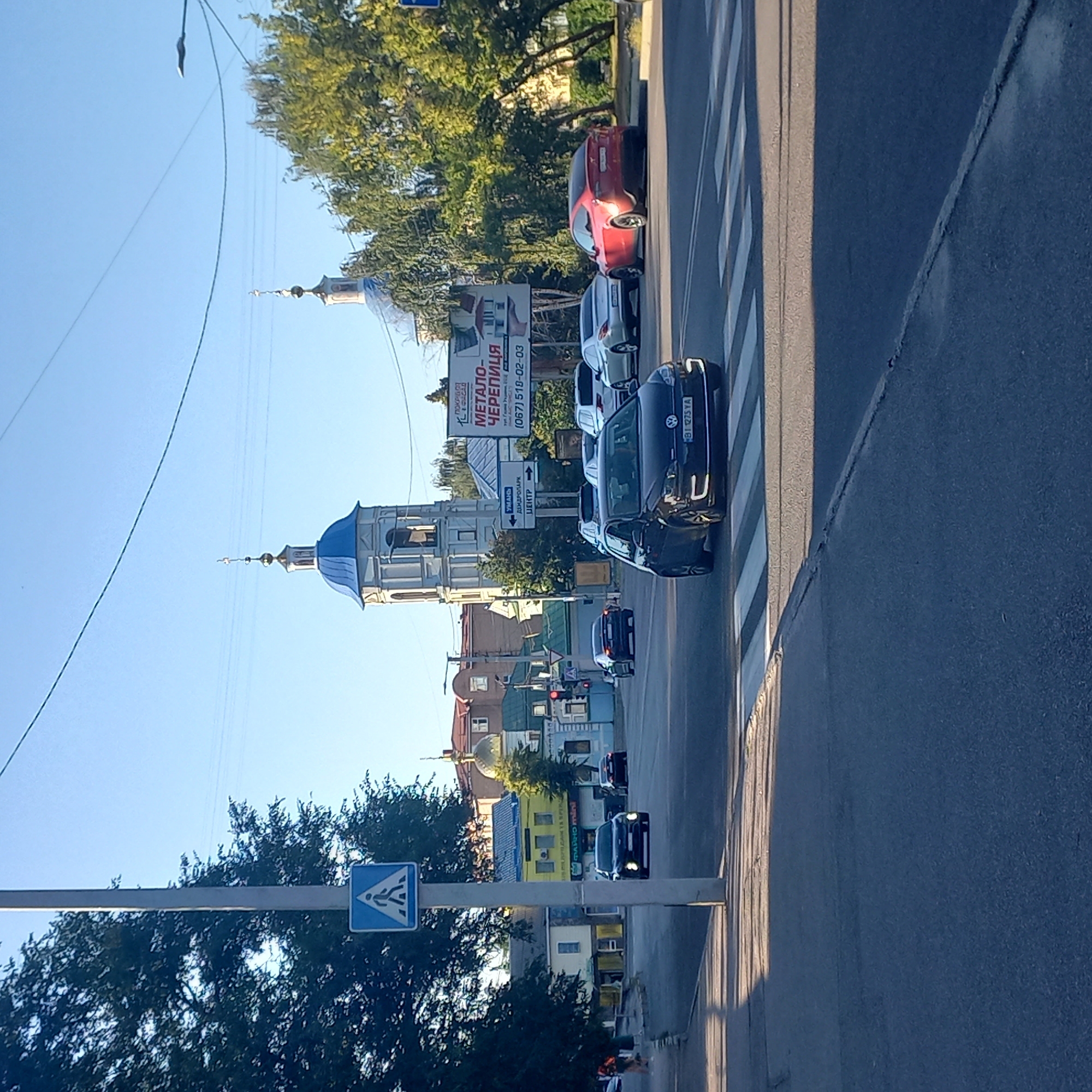 The image captures a street scene in Kropyvnytskyi, Ukraine, on a clear day, likely in the afternoon given the shadows. In the foreground, a dark Volkswagen Golf is stopped at a pedestrian crossing. The car has its headlights on, and its license plate is visible, reading "BI 1275 YA". To the right, a red Tesla Model X is parked on the side of the road. In the background, a multi-story building with a blue domed church tower is prominent. Another church with a golden dome is visible to the left of the blue-domed tower. A large billboard with Cyrillic text and an image of roofing materials is also present in the mid-ground. Several other vehicles are on the road, and trees line the street, casting shadows on the pavement. A pedestrian crossing sign is visible on a pole in the left foreground. The overall environment is that of a typical urban street with traffic and buildings.