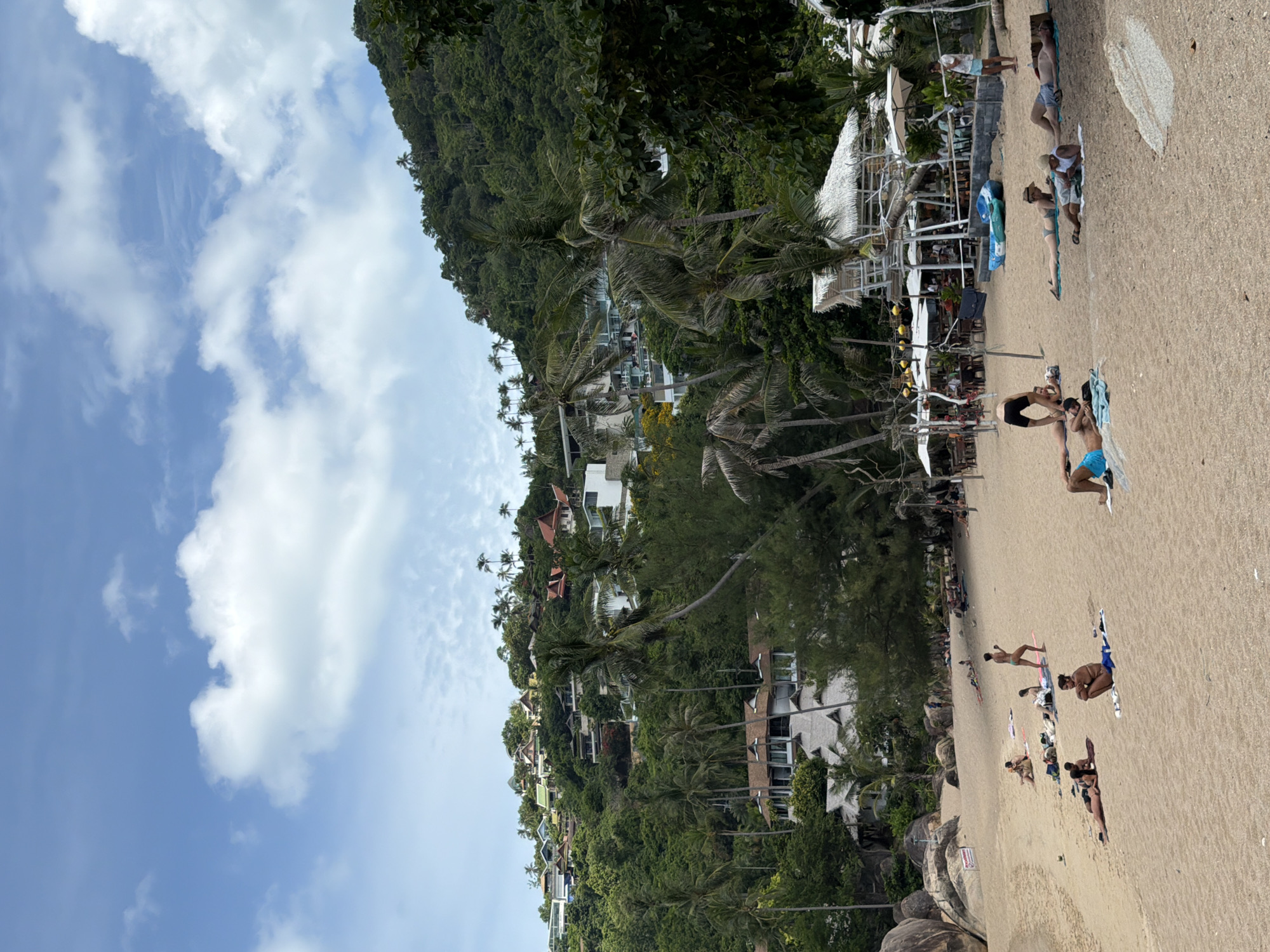 A wide shot of a sandy beach in Ko Samui District, Thailand, on a cloudy day. The beach is dotted with sunbathers, some in bikinis and swim trunks, lying on towels or sitting. A few palm trees line the beach, and in the background, a lush green hillside is covered with houses and more palm trees. A few beach umbrellas and a small hut-like structure are visible, suggesting a resort or restaurant area. The sky is a mix of blue and white clouds.