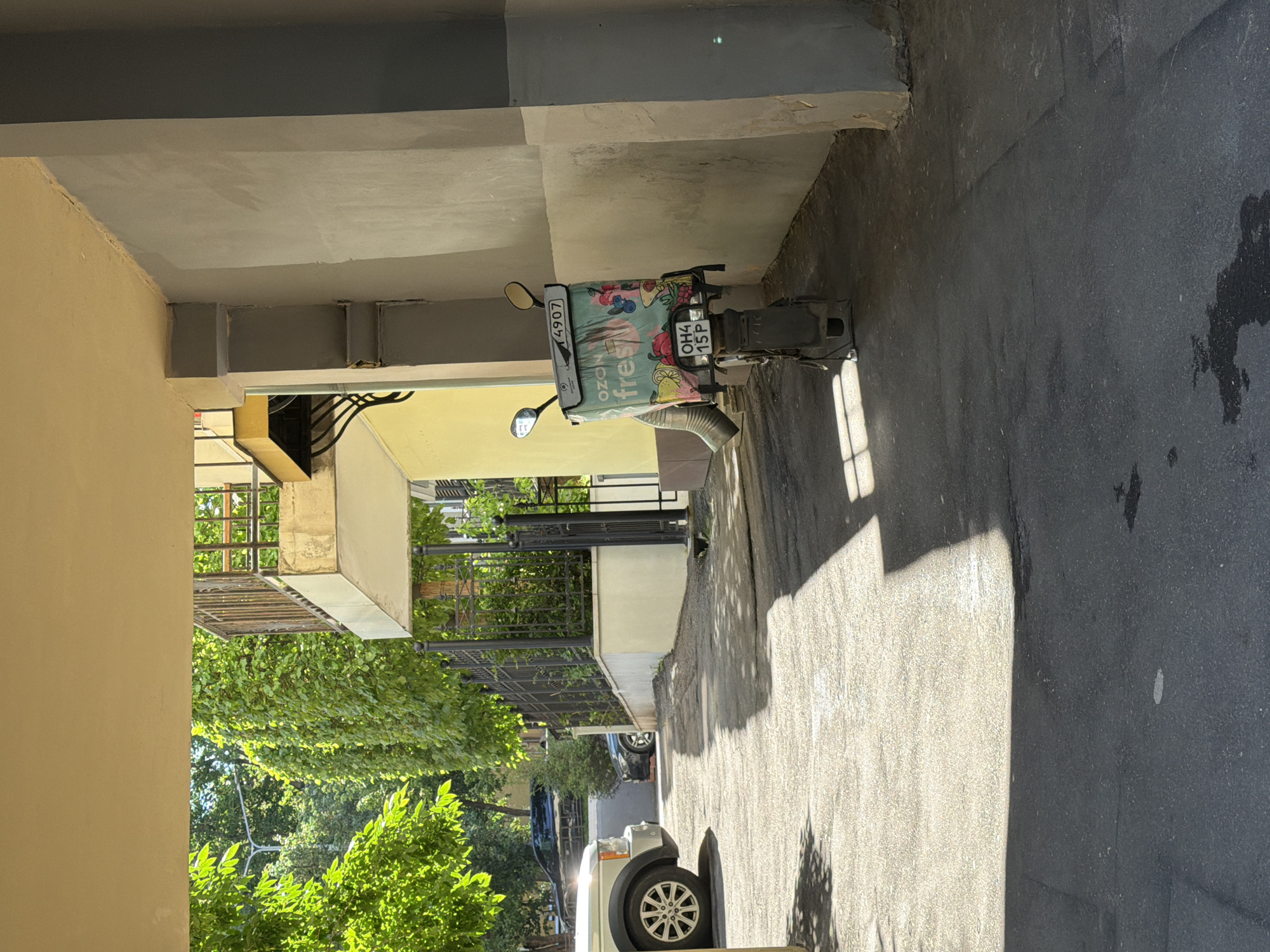 The image depicts a delivery scooter parked on an asphalt surface within what appears to be an urban courtyard or alleyway. The scooter is equipped with a large, insulated delivery bag on the rear, featuring the "Ozon fresh" logo and graphics of fruits and berries. A license plate with the characters "OH4 15P" is visible on the back of the scooter. In the background, a white SUV is parked, and a black car is further down the alley. Lush green foliage and trees line a black metal fence to the left, suggesting a well-maintained exterior space. The architecture includes a pale yellow building with a balcony and a light grey concrete wall on the right. Sunlight casts shadows across the ground, indicating daytime. There are no people visible in the frame. The scene is consistent with a delivery service operating in a residential or commercial area of Moscow, Russia.