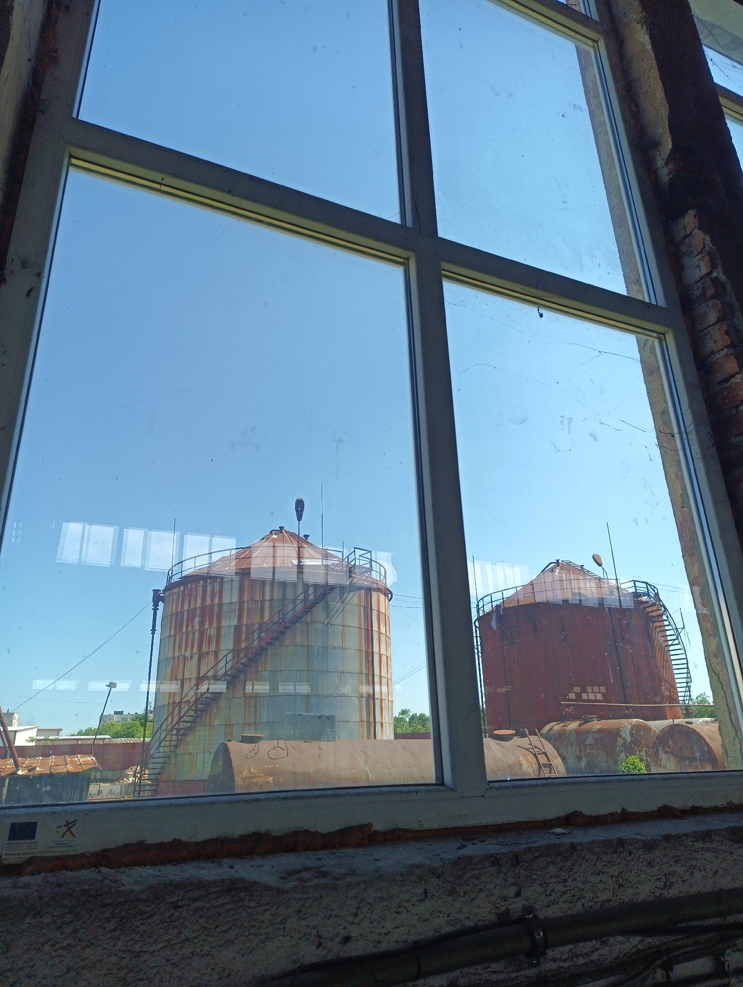 A slightly high-angle shot through a window shows two large metal tanks under a clear blue sky. The window is framed with white, and the sill is made of weathered, light-gray concrete. Brickwork is visible on the right side of the window frame.
The tanks are cylindrical with rounded tops and exhibit significant rust. The larger tank on the left is silver-gray with streaks of rust, while the one on the right is primarily red-brown with heavy rust. Each tank has a metal staircase leading to the top, and they are connected by a series of horizontal pipes.
In the distance, behind the tanks, is a line of green trees and the tops of buildings. The location is Unknown, Unknown.