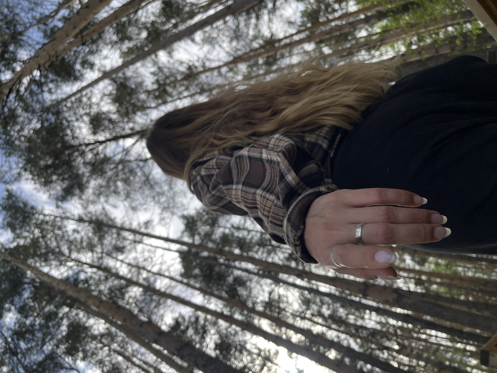 A low-angle shot looking up into a forest of tall, slender trees, likely pine or similar conifers. The sky is visible between the branches, appearing partly cloudy. In the foreground, a person is partially visible, wearing a brown and black plaid shirt, dark sweater and has long blonde hair. The person's left hand is prominent, adorned with two rings on the ring finger and index finger, respectively. The fingernails have a light pink color. The photo was taken in the city of Chaikovsky, Russia.