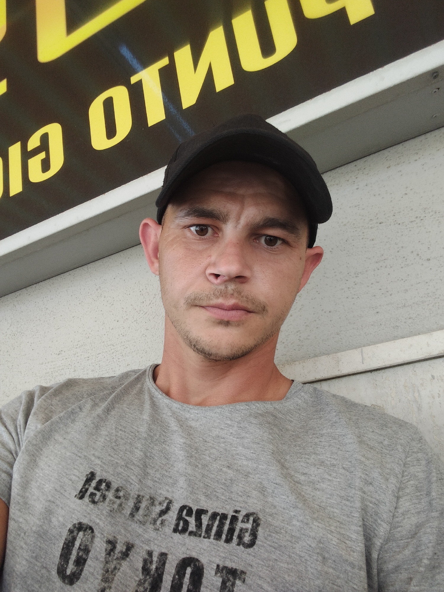 A close-up selfie shows a man wearing a black baseball cap and a grey t-shirt with text on it, standing outdoors. He has short brown hair, brown eyes, and a light stubble. Behind him, there is a textured wall and a sign with yellow lettering against a dark background. The sign appears to be a business sign with the text "PUNTO" and "CIAGIA". This business is located in the city of Mondragone, Italy.