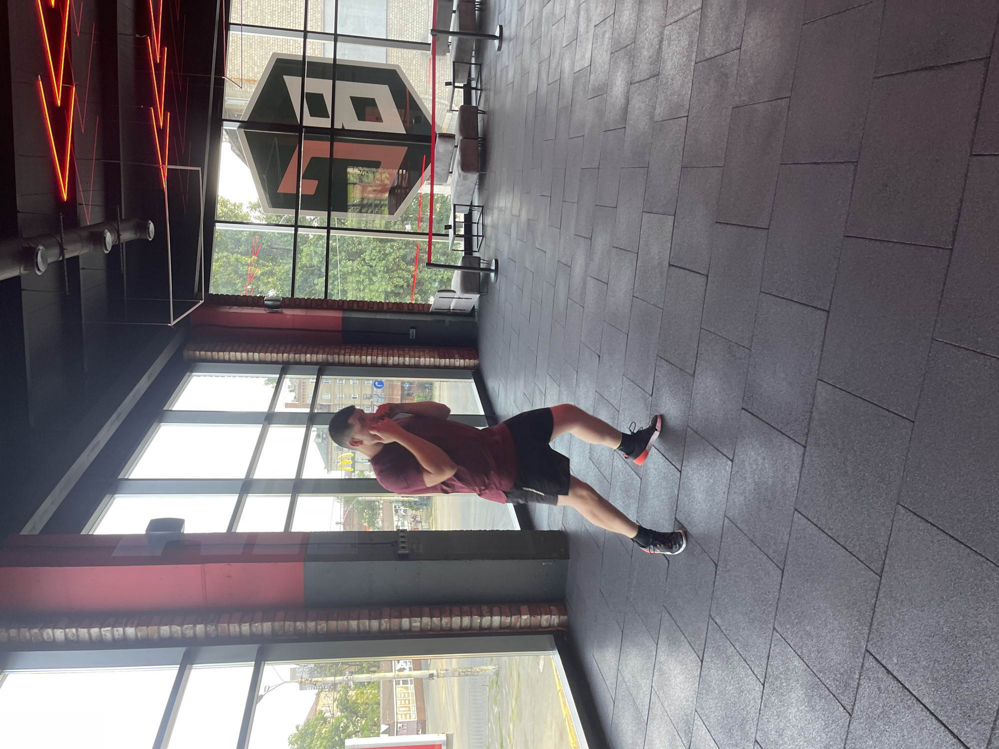 An athletic young man with short, dark hair is shown in a boxing stance within a modern gym. He is wearing a maroon t-shirt and black shorts with a white stripe and black athletic shoes with orange accents. His hands are raised in front of his face, and his body is positioned as if he is about to throw a punch or is defending himself. The gym features large windows with a view of an urban street and trees, a brick pillar, and a black ceiling with red neon lighting. The floor is covered with dark grey, interlocking rubber tiles. In the background, through the window, a large building with a distinctive logo featuring a stylized "HB" is visible. Several modern grey couches and tables are arranged in a waiting area behind a red velvet rope barrier.