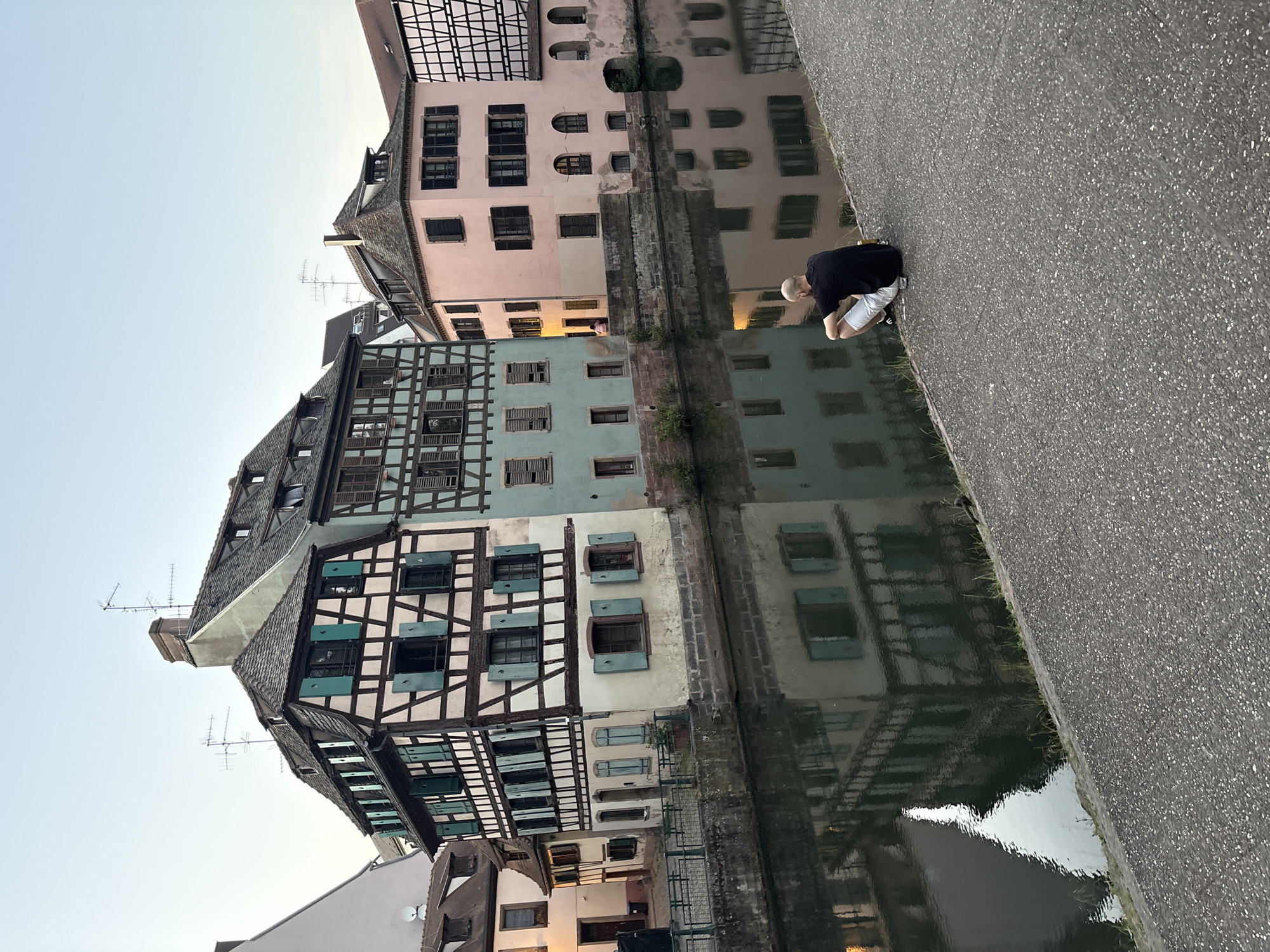 A man is crouching on a paved road next to a canal that reflects timbered buildings in Strasbourg, France. He is wearing a black shirt and light-colored shorts or pants. The buildings vary in color with light blue, pink, and cream facades. The canal water is still, creating a clear reflection of the buildings. The sky is light, indicating it could be early morning or late evening.