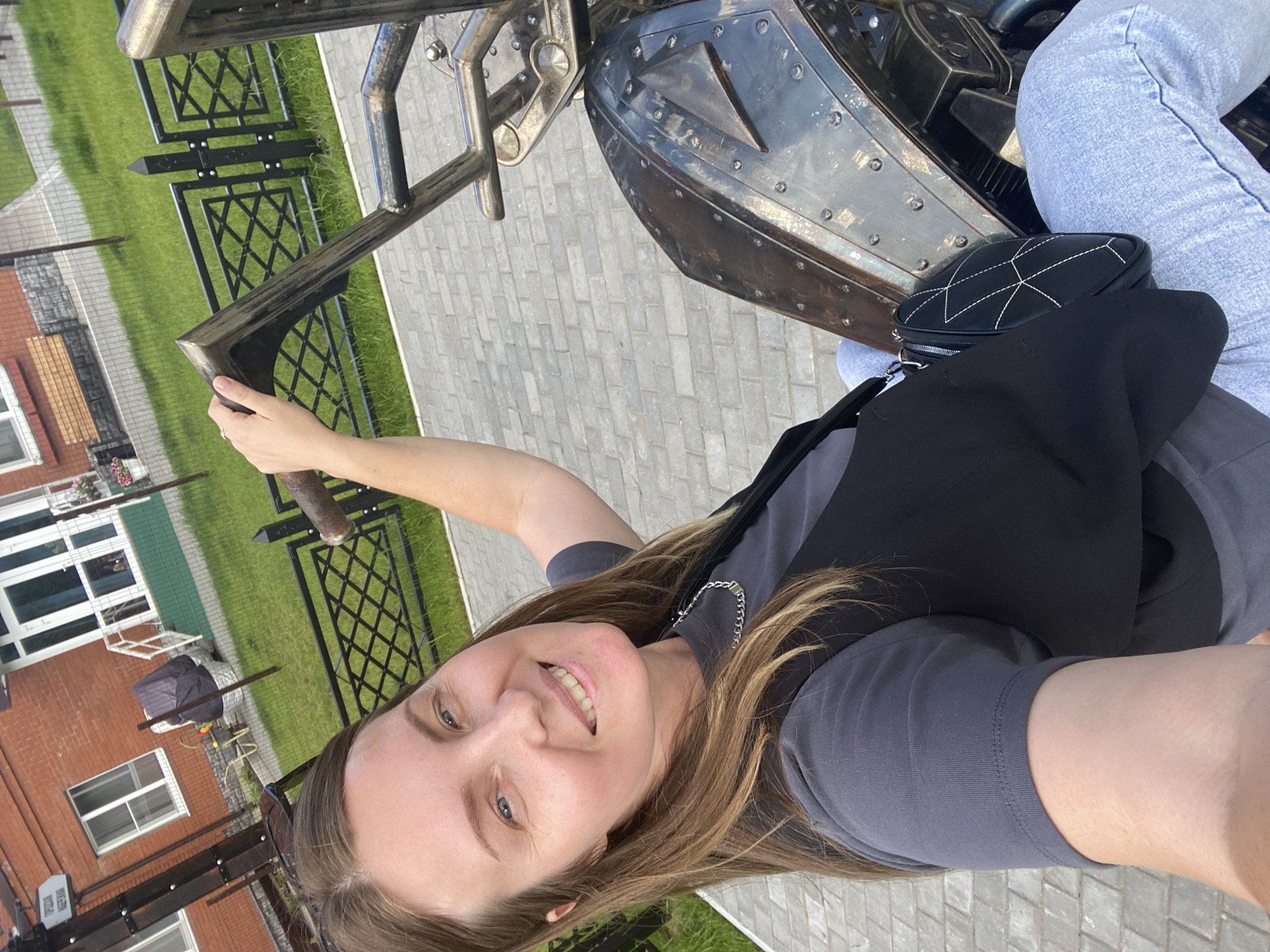 This is a selfie taken by a woman in Chaikovsky, Russia. She is smiling and looking at the camera, with her right arm extended, holding onto a metal railing. She is wearing a grey t-shirt, a black vest, and light blue jeans. A small black quilted purse with white stitching is visible on her lap.
The setting appears to be outdoors, possibly a park or a decorative area. To her left, there is a brick building with white-framed windows. Behind her, a green lawn is visible, bordered by a black metal fence with a decorative diamond pattern. The ground is paved with grey interlocking bricks.
The lighting suggests it is daytime, likely with clear weather. The woman's expression is happy and relaxed, indicating a pleasant mood. The metal object she is holding onto has a dark, metallic, possibly steampunk-inspired design, with rivets and decorative elements.