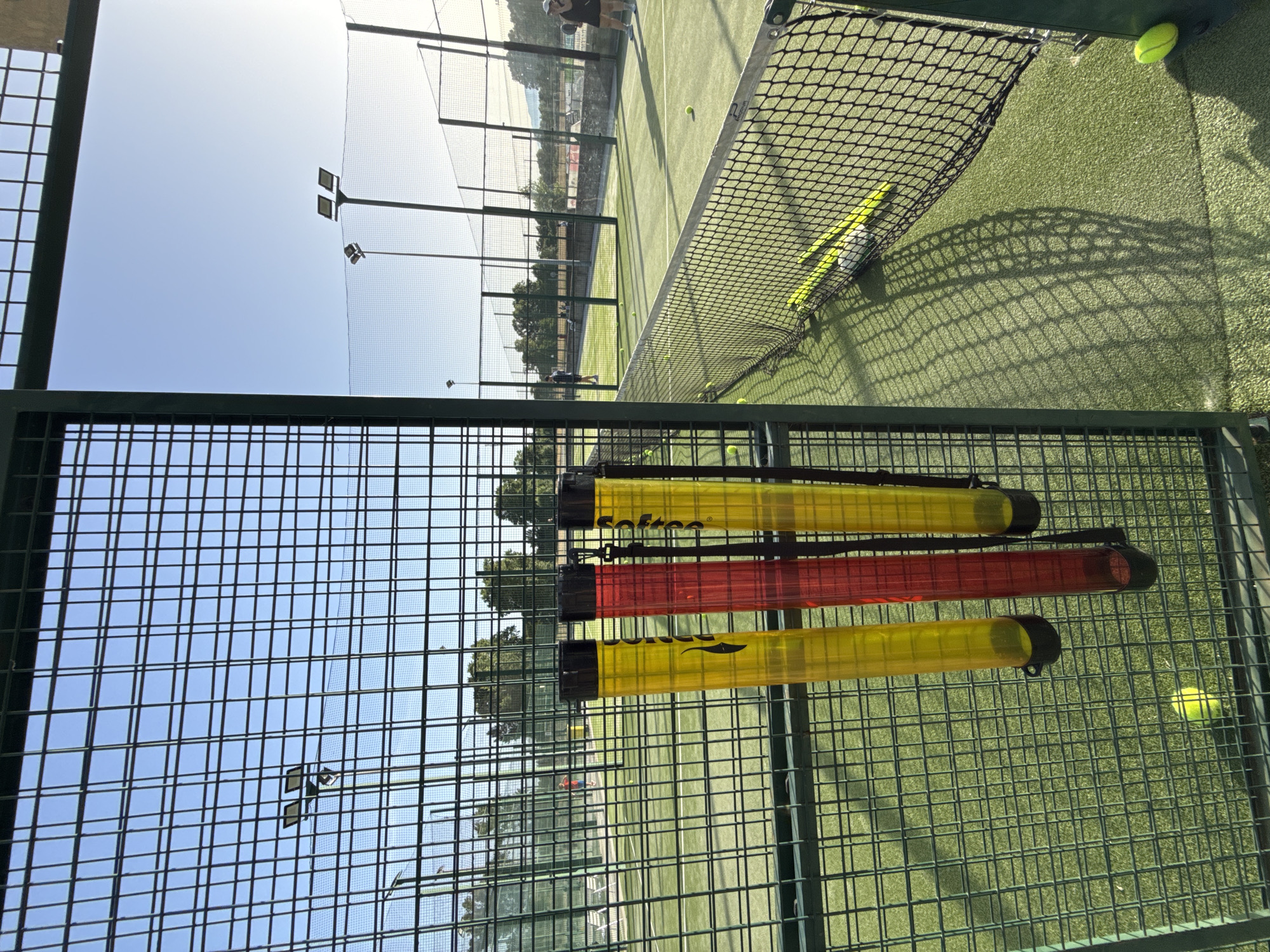 The image shows an outdoor padel tennis court in Vidreres, Spain. The court is made of green artificial turf and is enclosed by a metal fence. A net divides the court in half. Three transparent tubes, two yellow and one red, filled with padel balls are attached to the fence on the left side of the image. Several yellow padel balls are visible on the court. In the background, there are more padel courts, trees, and a clear blue sky with the sun setting, casting long shadows across the court. Two people are visible on the courts; one is standing near the net on the right, and another is playing on a court further in the background. There are also some stadium lights visible above the courts.
