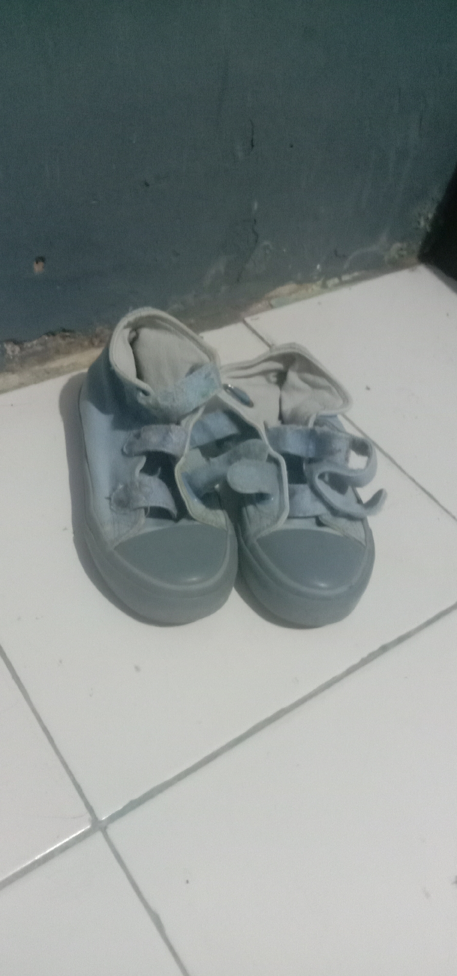A pair of light blue high-top canvas sneakers with gray rubber soles and white laces is placed on a white tiled floor. The sneakers are positioned side-by-side, with their tongues pulled out and the laces loosened. The background consists of a dark gray wall. The scene appears to be an indoor setting, likely a home or residential area, given the presence of floor tiles and a simple wall. The image is taken from a slightly elevated angle, looking down at the shoes. The lighting suggests an artificial light source, possibly a flash, illuminating the sneakers and casting subtle shadows. The sneakers appear to be children's shoes due to their size and style. There is no indication of any specific location in Kendari, Indonesia, other than it being an indoor environment.