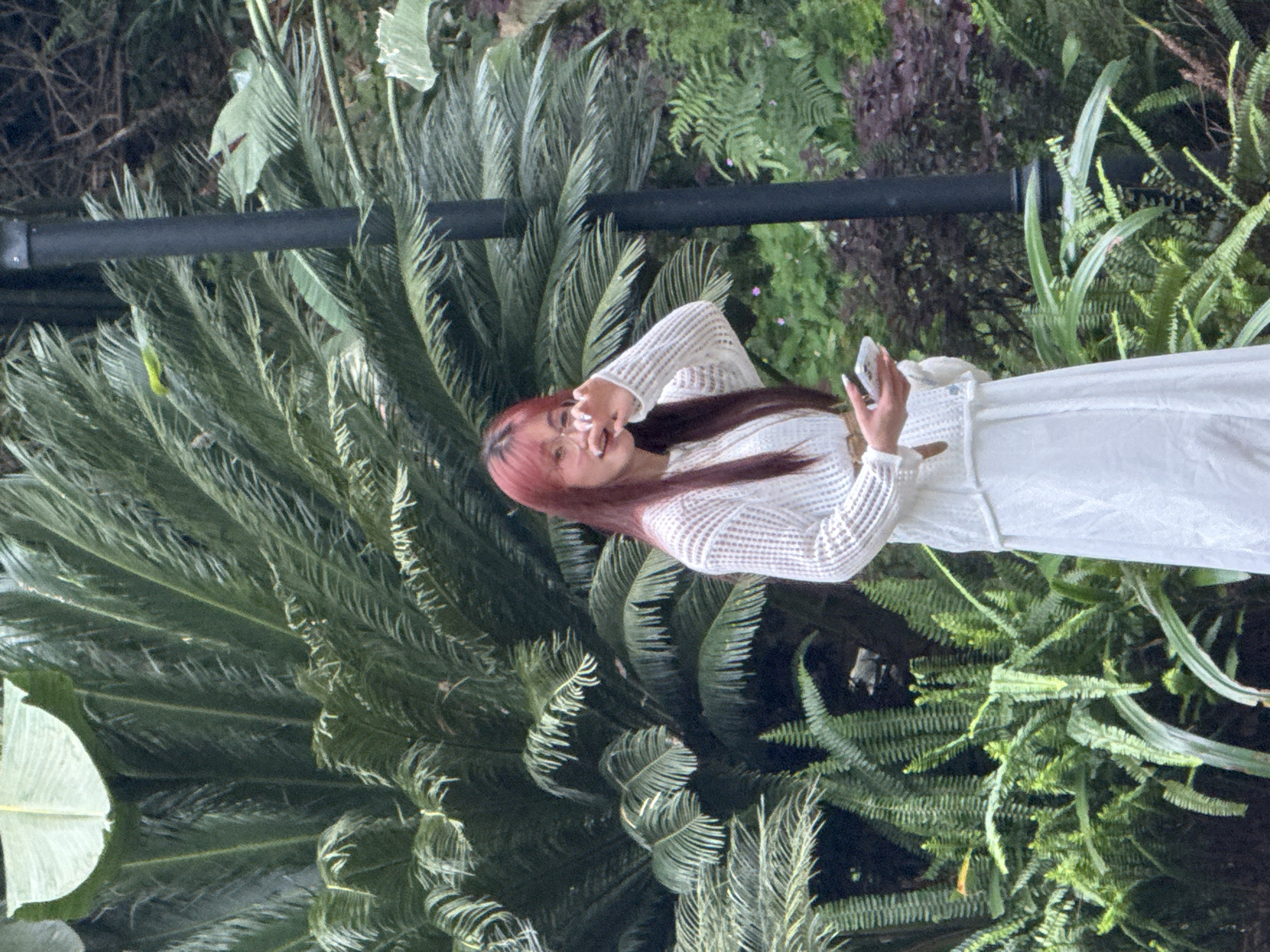 A woman with pink-tinted hair, wearing a white sweater and a white skirt, stands amongst dense greenery. She is holding a mobile phone in one hand and making a gesture with the other. The background consists of various plants, including ferns and cycads. The image was taken in Jiangjin, China.