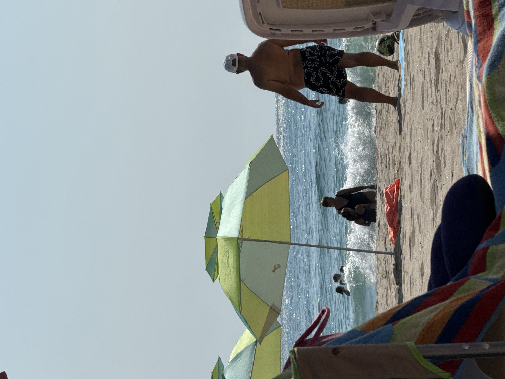 The image depicts a sunlit beach scene with the Black Sea in the background. In the foreground, a colorful striped towel is draped over a chair. Two beach umbrellas, one predominantly green and yellow and another partially visible with blue and green hues, are set up on the sand. In the middle ground, several people are visible in the shallow water. One person, a man wearing a white cap and patterned swim shorts, stands facing away from the camera, looking towards the sea. Further out, other individuals are wading or swimming. The water is calm with small waves breaking on the shore. The sandy beach is littered with a few items, including a folded orange towel. The sky is clear and a pale blue. The scene is characteristic of a summer day at a coastal resort, possibly in the vicinity of Obrochishte, Bulgaria, known for its proximity to beaches.