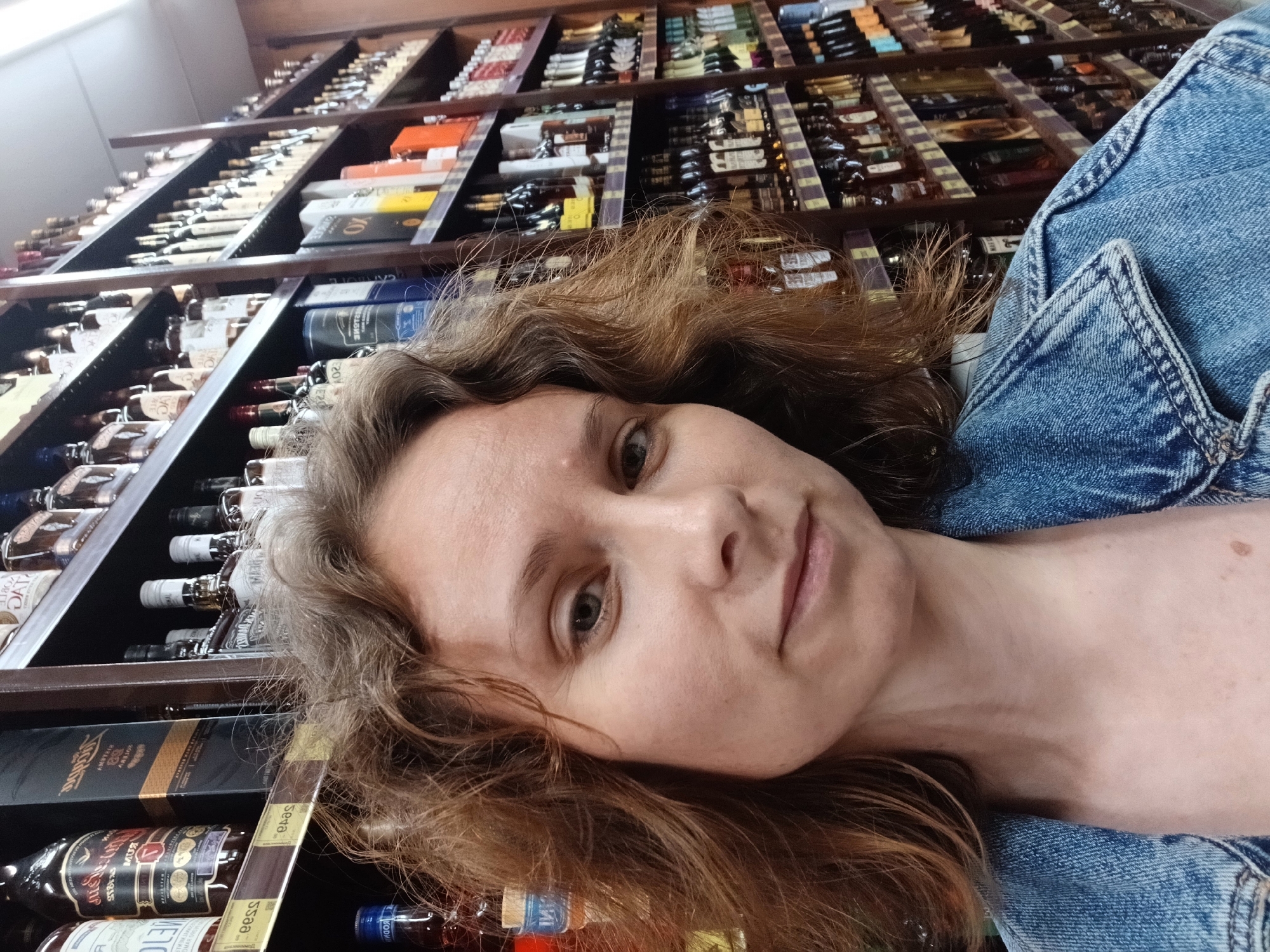 The image is a selfie taken by a woman with brown curly hair and fair skin. She is wearing a blue denim jacket. The background shows shelves stocked with bottles of alcohol, suggesting she is in a liquor store or a similar establishment. The lighting is somewhat dim, with reflections visible on the bottles. The overall composition focuses on the woman's face and upper body, with the store's inventory filling the background. The location is Yekaterinburg, Russia.