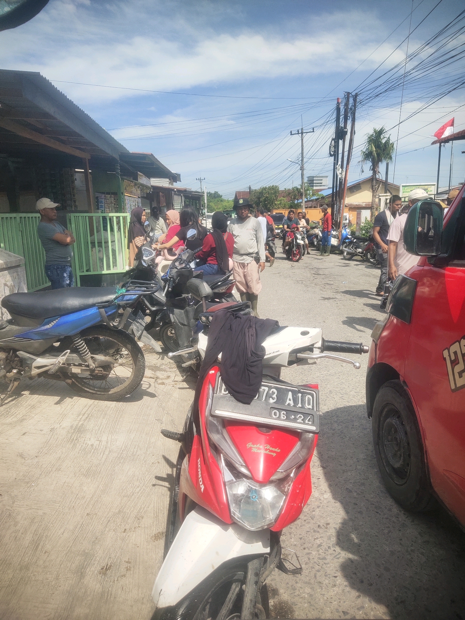The image depicts a street scene in the city of Medan, Indonesia, on a sunny day with scattered clouds. The foreground is dominated by the front of a red and white motorcycle, with its license plate reading "73 AIQ 06-24." To the right, a portion of a red vehicle, possibly a car or van, is visible, showing the number "12" on its side. The middle ground features several parked motorcycles and a group of people, some of whom are conversing or standing around. A man in a light-colored cap and a gray t-shirt is standing with his arms crossed in front of a building with a green metal fence. Other individuals are seen in the background, some on motorcycles and others walking. The environment appears to be a commercial or residential street, with buildings lining one side and power lines overhead. The road surface is asphalt with some visible patches and debris. A palm tree is visible in the distance.