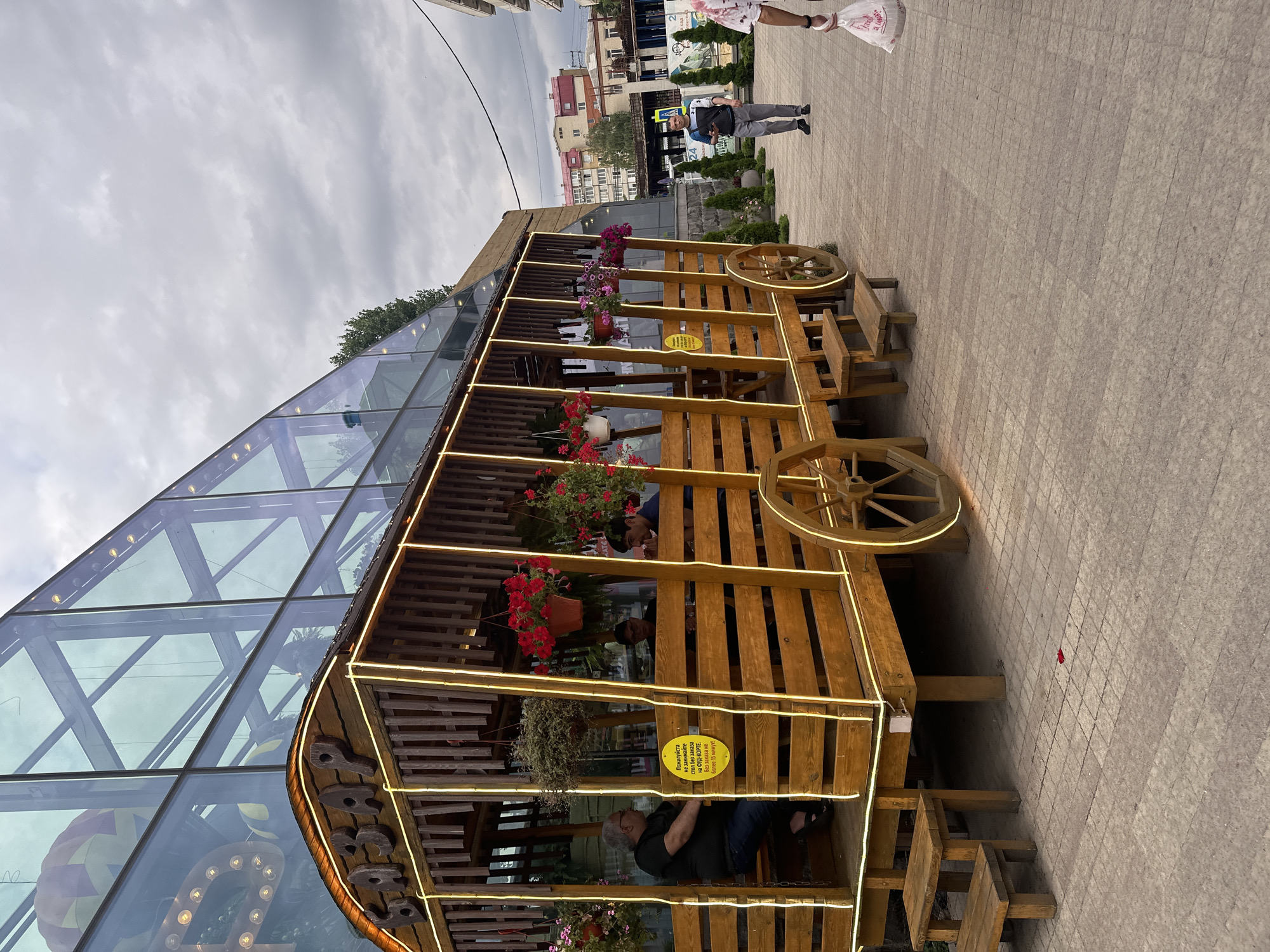 The image shows a rustic wooden structure resembling a large wagon or carriage, decorated with bright yellow string lights, positioned on a paved walkway in front of a modern building with a glass facade. This establishment, identified by Cyrillic lettering on the wagon's roof that reads "БАZАР", is located in Stavropol, Russia. Inside the wagon, several people are seated at tables, engaged in conversation or dining. One man in a black t-shirt and jeans is seated on a bench facing away from the camera. Two other men are seated further inside the wagon. In front of the wagon, a man in grey pants and a dark jacket walks away from the camera, holding a tablet. To the right, a person in a white shirt with a red pattern is walking towards the camera, carrying a white plastic bag. The background shows a cloudy sky and buildings in the distance. The overall scene depicts a casual outdoor dining or market area.