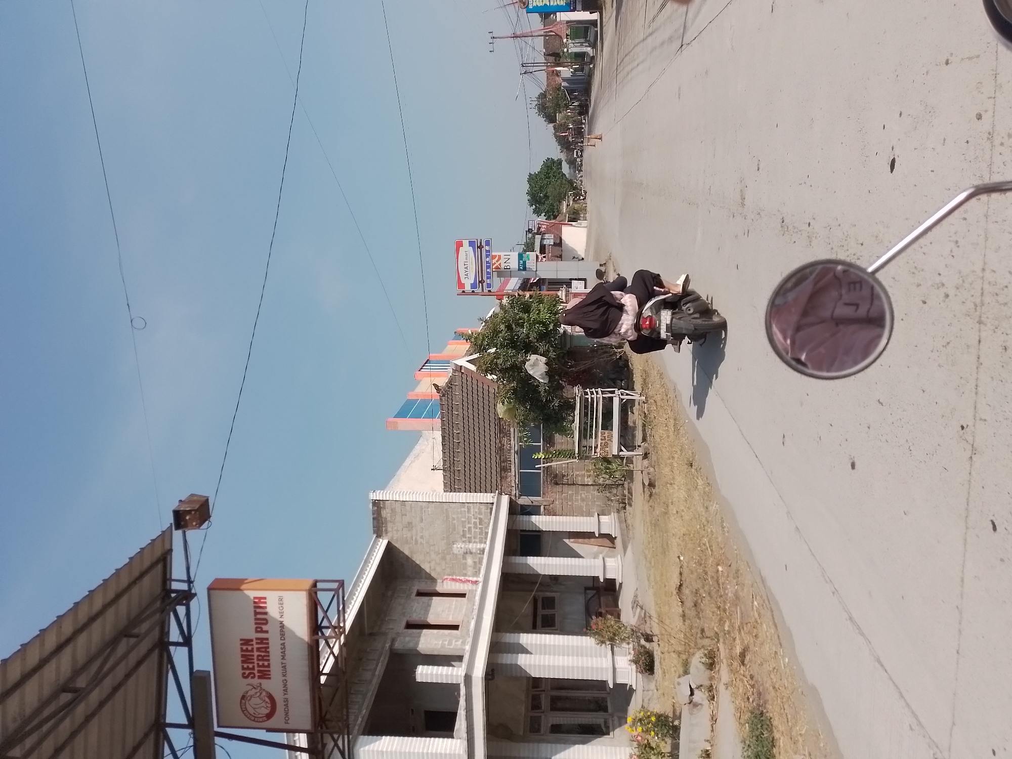 This is a medium-shot, eye-level, outdoor, daytime photograph taken from a motorcycle in Ngancar, Indonesia. The scene depicts a rural or suburban road with buildings and vegetation on either side. In the foreground, a motorcycle with a rider wearing a black hijab and a pink and white patterned jacket is visible, heading away from the camera. The motorcycle's rearview mirror, which reflects a pink and white patterned shirt, is also in the foreground. To the left of the road, there is a partially constructed building with white columns and a tiled roof, and a sign with "SEMEN MERAH PUTIH" and "FONDASI YANG KUAT MASA DEPAN NEGERI" printed on it. Further down the road, there are more buildings, including one with a blue and orange facade, and signs for "JAYATImart" and "BNI ATM". The sky is clear and blue, indicating a sunny day. The road itself is paved and appears to be in good condition, with some cracks and debris. The environment suggests a commercial or residential area in a relatively undeveloped part of the city.