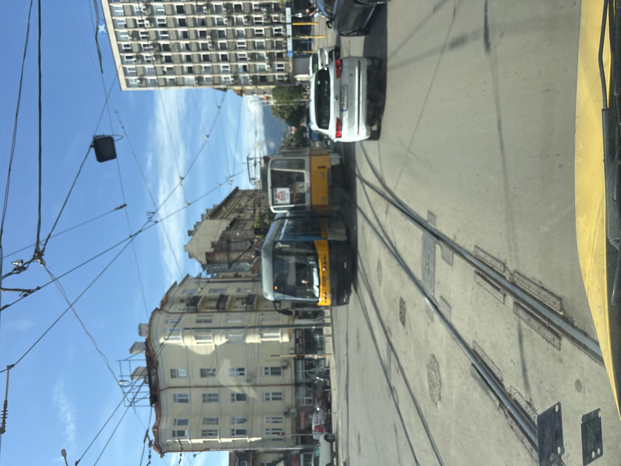 This is a daytime, eye-level, medium-shot, slightly blurred photograph of a street scene in Sofia, Bulgaria, likely taken from a vehicle. The foreground is dominated by the yellow hood of a car, with a portion of a windshield wiper visible. Tram tracks are prominent, stretching across the asphalt road.
In the middle ground, two trams are positioned on the tracks. The tram on the left is blue and yellow, with its front facing the camera and slightly to the right. The tram on the right is yellow and white, positioned further down the tracks and facing away from the camera. The front of the yellow tram has a sign.
Several vehicles are visible in the background. A white SUV is in the right lane, facing away from the camera. Behind it, another white vehicle is parked. To the left of the trams, other cars are parked on the side of the road.
The urban environment includes several buildings. On the left, a light yellow, ornate building with several floors and windows is visible. On the right, a tall, grey, modern building with many windows stretches upwards. Overhead, a complex network of tram wires and support poles crisscrosses the blue sky, which has scattered white clouds. The overall impression is of a busy urban intersection with public transportation.