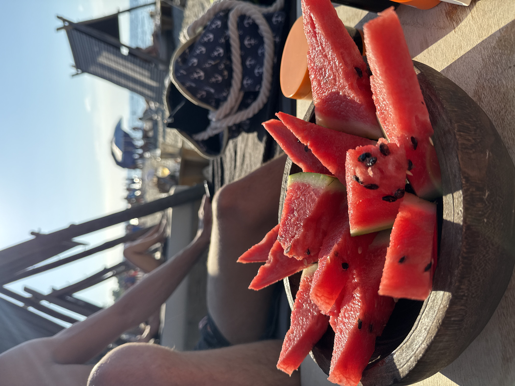 The image shows a close-up of a bowl of watermelon slices, positioned on a wooden surface. The watermelon is cut into thin, triangular wedges, with visible black seeds. The bowl itself is made of dark wood and has a rustic texture. In the background, the scene suggests a beach or outdoor setting, with hints of a wooden structure, the sea, and a clear sky, indicating a sunny day, likely in the late afternoon given the warm light and long shadows. A person's legs and arm are partially visible in the lower left and center of the frame, suggesting they are seated and enjoying the watermelon. Behind the bowl, a navy blue bag with a white rope and anchor pattern is visible, along with an orange object. The overall atmosphere is relaxed and summery. The location context provided indicates Pallini Municipal Unit, Greece.