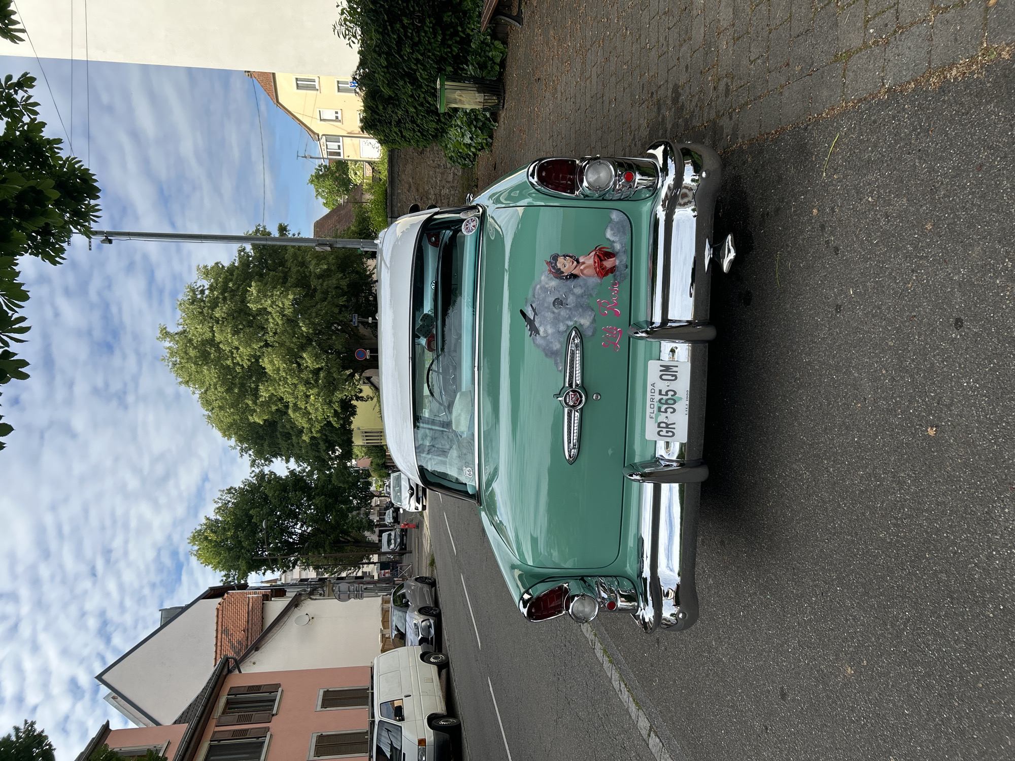 The image shows the rear of a vintage green and white car parked on a street in Strasbourg, France. The car has a Florida license plate that reads "GR-565-QM." A mural of a woman and an airplane decorates the car's trunk. The street is lined with buildings of varying colors and architectural styles. Several other parked vehicles can be seen further down the street. Trees and foliage are visible along the street. The sky is partly cloudy. The road surface is asphalt.