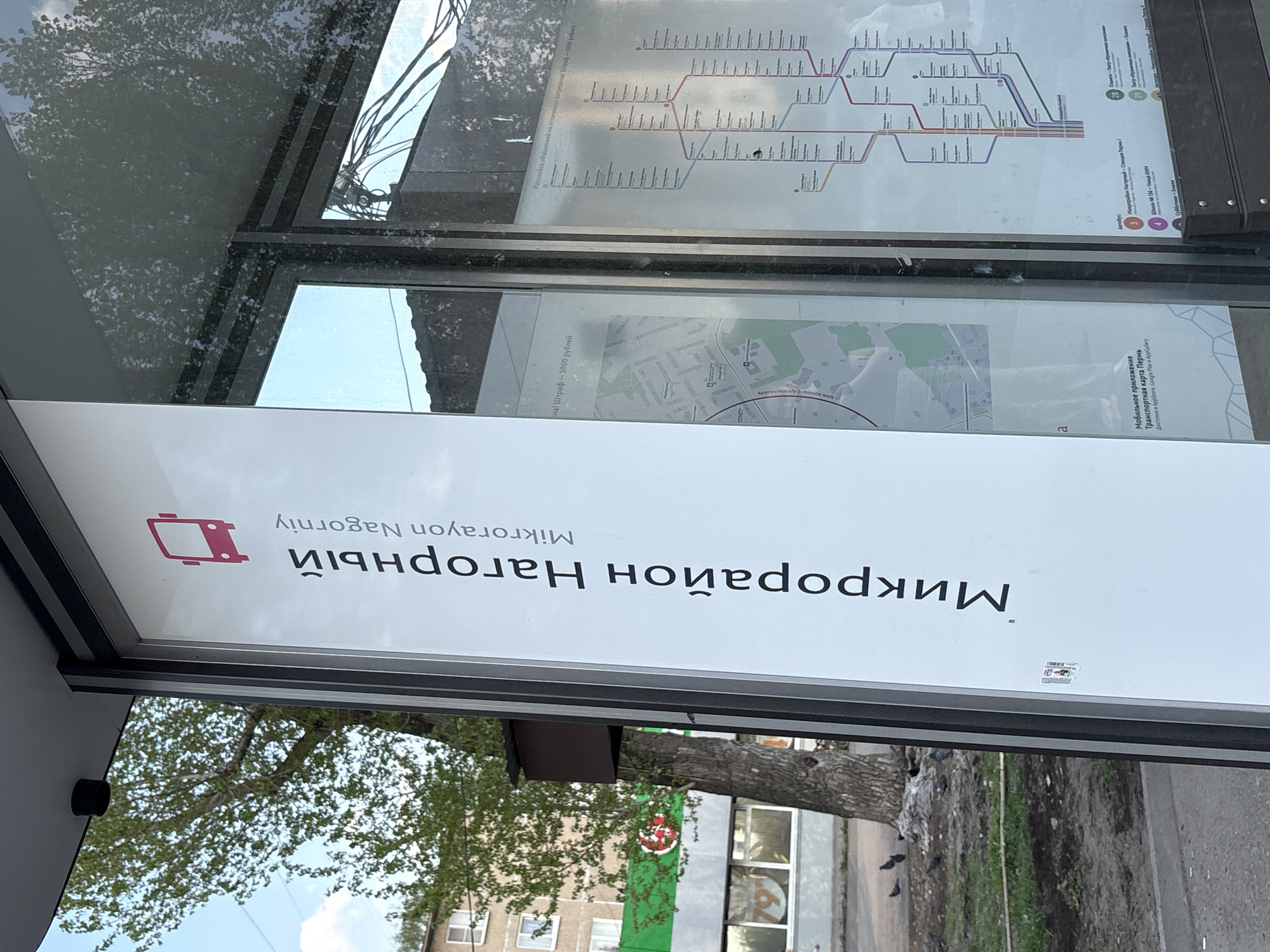 The image shows a bus stop in Perm, Russia, specifically in the Nagorny microdistrict (Микрорайон Нагорный). A sign on the bus stop indicates the name of the microdistrict in both Russian and English (Mikrorayon Nagorniy). Above the sign is a map of the city's transportation routes. A smaller map displays the surrounding area with text reading "approximately 5-minute walk". Adjacent is a QR code and text that reads "Mobile application Transport card Perm". Further down, the text says "Available in AppStore, Google Play and AppGallery". The text below that indicates that bus number 3 goes to the Nagorny microdistrict, the Perka Station, School number 106, and towards the street named Ulitsa KIM. Bus number 4 goes to Subbotina-Khmeli. The text reads "Sticking announcements on stops is forbidden! Fine - 5000 rubles!". The image also shows the surrounding environment, including trees, a building, and a green pole.