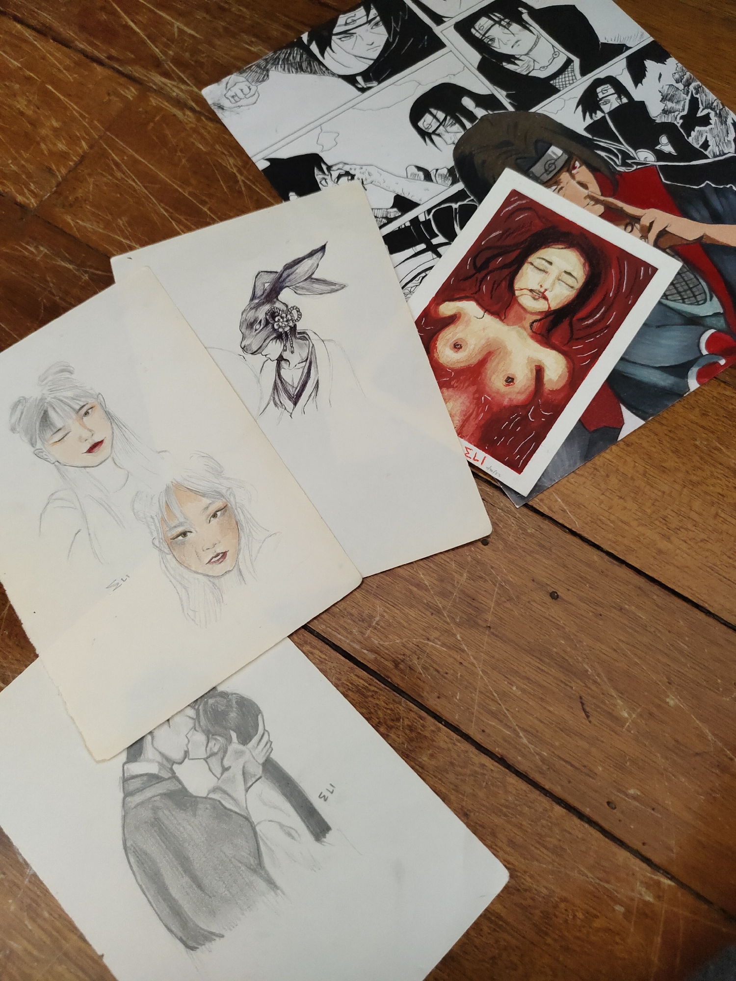 The image displays several pieces of artwork on a wooden surface, likely a table or floor, with visible wood grain and scratches. The artworks are primarily sketches and a painting.
One piece, a painting, depicts a person lying down with their upper body exposed, in what appears to be a pool of red liquid. This artwork is positioned towards the right side of the frame, partially overlapping other pieces.
Another artwork, a black and white drawing with some colored elements, shows characters from the anime series Naruto, specifically Itachi Uchiha. This piece is visible at the top and right of the frame.
To the left of the painting and Naruto artwork, there are several sketches on paper. One sketch shows a person wearing a rabbit-like mask with decorative elements. Below this, there are sketches of two female faces with stylized hair and makeup. A sketch of a couple kissing is visible at the bottom of the frame, with one person's hand gently touching the other's face.
The lighting suggests an indoor setting. The overall scene is an arrangement of creative works on a textured wooden background. No specific location in Davao City, Philippines, can be determined from the visual information provided.