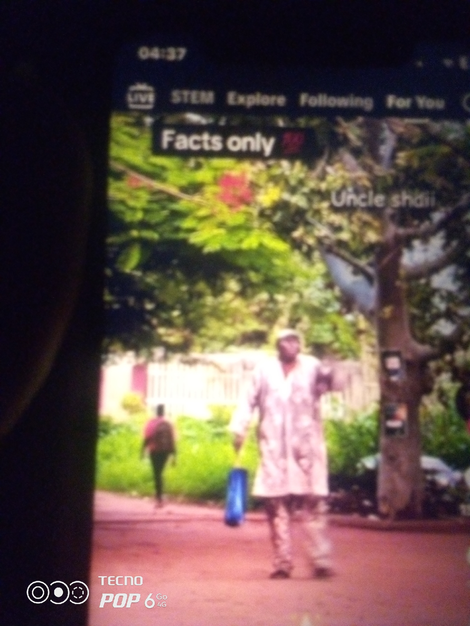 The image is a screen capture from a mobile device showing a video stream. The top of the screen displays a time of "04:37" and navigation options including "LIVE", "STEM", "Explore", "Following", and "For You". Prominently displayed is a text overlay "Facts only" with a small red icon next to it.
The video content itself shows an outdoor scene, possibly a park or a street in a city. A person, appearing to be a man, is walking towards the camera, dressed in a light-colored, long garment and pants. He is holding a blue object, possibly a bag, in his left hand. To the left of this central figure, another person is walking away from the camera, wearing a dark jacket over a pink shirt and dark pants. In the background, there are trees with green foliage and some pink flowers. There is also a fence visible in the mid-ground.
The bottom left corner of the screen displays a "TECNO POP 6 4G" logo.
The overall image quality is somewhat blurry and the lighting suggests it might be daytime. The scene does not provide specific visual cues to pinpoint the location to Howayi, Nigeria.