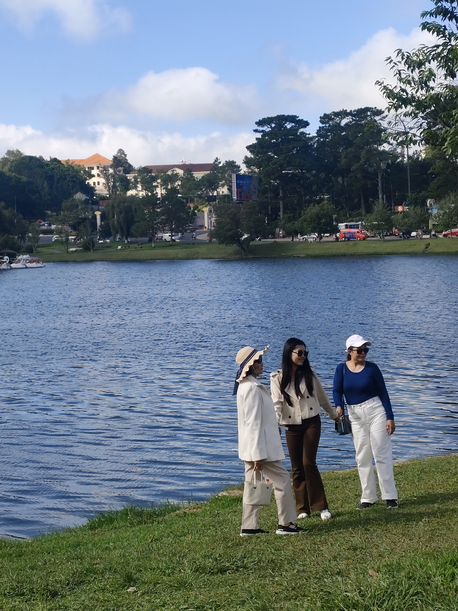 Three individuals stand on a grassy bank overlooking a wide, calm body of water, likely Xuan Huong Lake in Da Lat, Vietnam. The person on the left, wearing a wide-brimmed hat, a cream-colored coat, and cream pants, holds a light-colored handbag. To their right, a person with long dark hair, sunglasses, a beige jacket over a striped top, and brown pants, holds hands with the individual on the far right. The person on the far right, wearing a white baseball cap, a dark blue long-sleeved shirt, and white pants, also holds a dark handbag. In the background, across the lake, are buildings with orange roofs, dense green trees, and a bright blue sky with scattered white clouds. Vehicles, including a red bus, are visible on a road running along the distant shoreline. The overall impression is of a leisurely day at a scenic location.