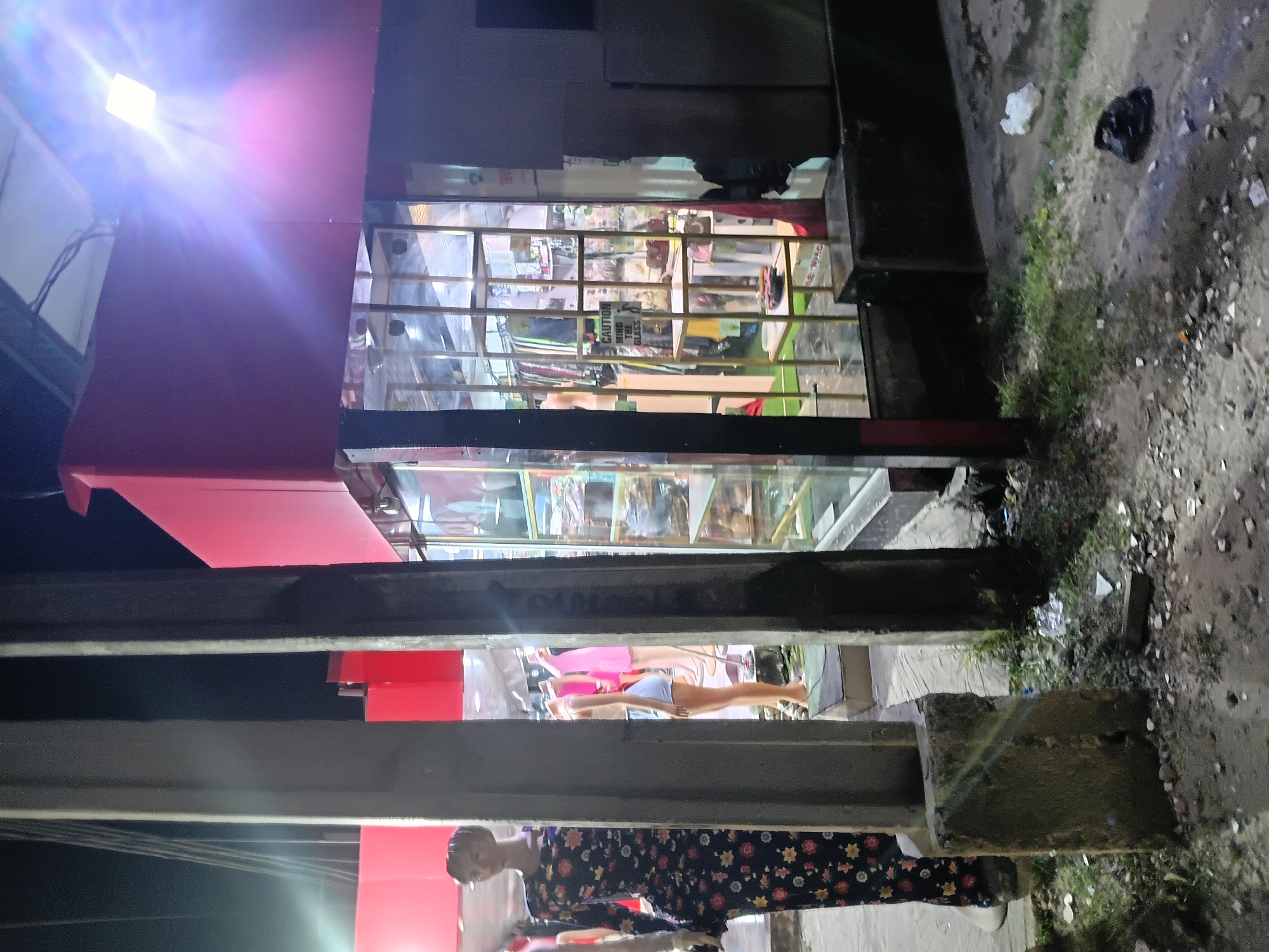 In Ado, Nigeria, this image captures a street view at night, likely a commercial area, with a prominent clothing store and mannequins. The store features a brightly lit interior with racks of clothing visible through a glass facade. A mannequin dressed in a long, floral-patterned dress stands in the foreground, partially obscured by concrete pillars. Another mannequin, wearing shorts and a top, is visible inside the store. The street outside is unpaved and littered with debris, including a plastic bag and what appears to be discarded building materials. A bright overhead light illuminates the scene, casting lens flares and creating a stark contrast between light and shadow. The overall impression is one of a bustling yet somewhat neglected urban environment.