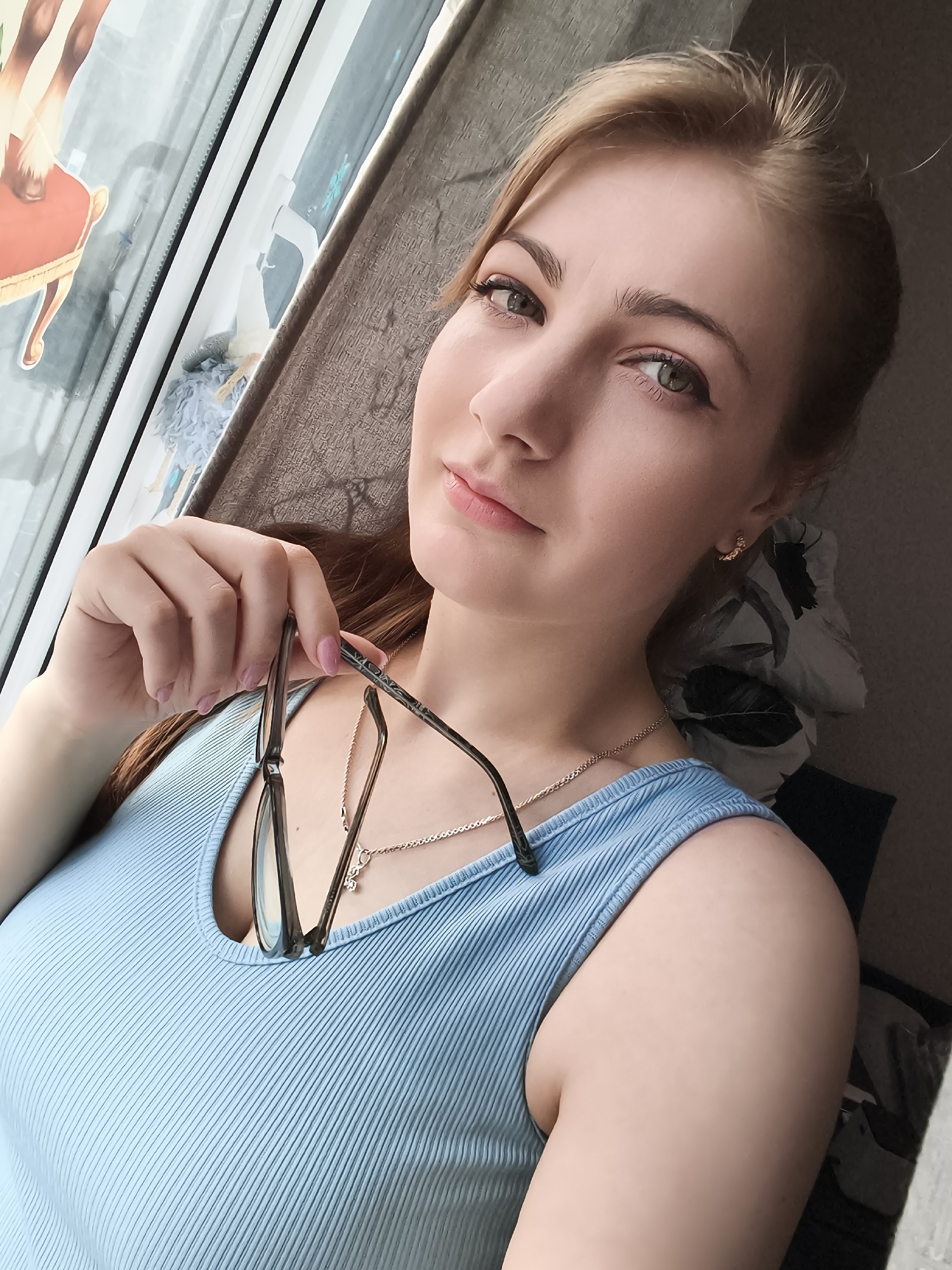 A young woman with blonde hair and green eyes is captured in a selfie, holding a pair of glasses in her right hand. She is wearing a light blue ribbed tank top and a silver necklace with a charm. Her left hand is resting on her chest, and her gaze is directed at the camera. The background suggests an indoor setting, possibly near a window with a curtain and a glimpse of a sticker on the glass. The lighting is natural, creating soft shadows on her face and body.