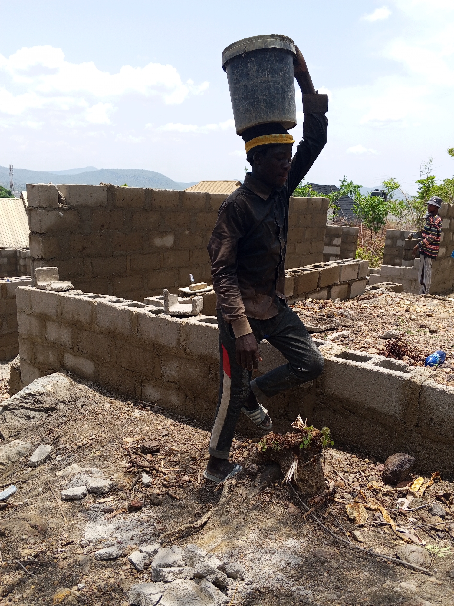 The image depicts a construction site in Lokoja, Nigeria. An adult male, dressed in a long-sleeved shirt and trousers with a red stripe, carries a large bucket on his head. He wears a yellow pad on his head for cushioning. The site features partially constructed brick walls. Another man in a striped shirt and hat stands nearby. The ground is littered with debris. In the background, buildings and hills are visible under a partly cloudy sky.