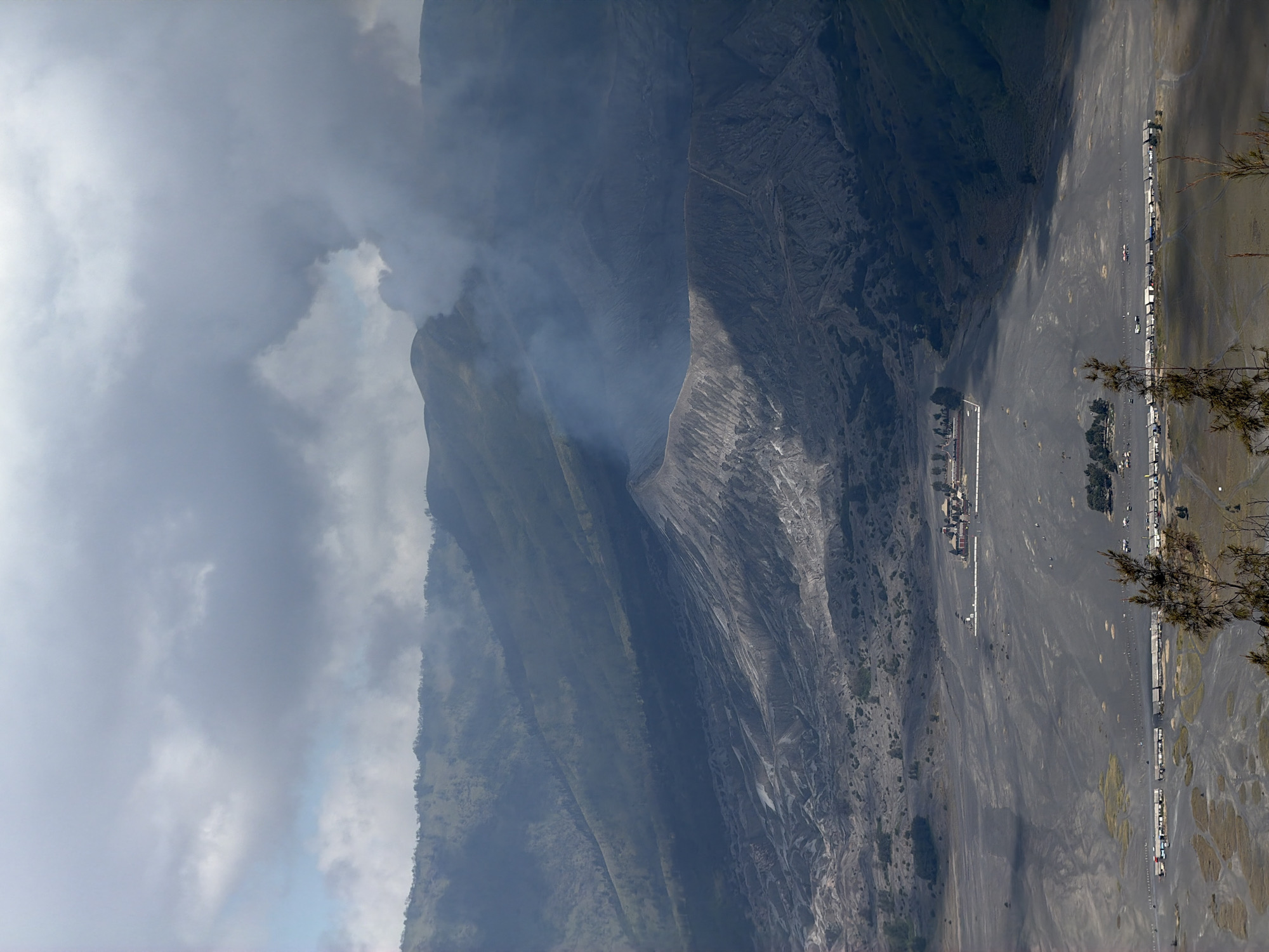 The image captures a panoramic view of Mount Bromo, a volcano in East Java, Indonesia, specifically looking towards Cemorolawang. In the foreground, a vast expanse of dark volcanic sand stretches out, punctuated by patches of dry grass and sparse vegetation. Several structures are visible in the distance; a long, low building complex with numerous small stalls or shops lines the bottom left of the frame, some with umbrellas erected outside. Further back, near the center of the image, a more substantial, multi-building compound with red-tiled roofs sits on the volcanic plain.
The middle ground is dominated by the rugged, ashen slopes of Mount Bromo, with its caldera clearly visible. Wisps of white smoke or steam emanate from the volcano's crater, indicating ongoing geothermal activity. The background is comprised of rolling, green hills and mountains, their slopes covered in vegetation and trees. The sky above is partly cloudy, with dramatic grey clouds casting shadows across the landscape. The overall impression is one of raw, natural power and a stark, volcanic environment.