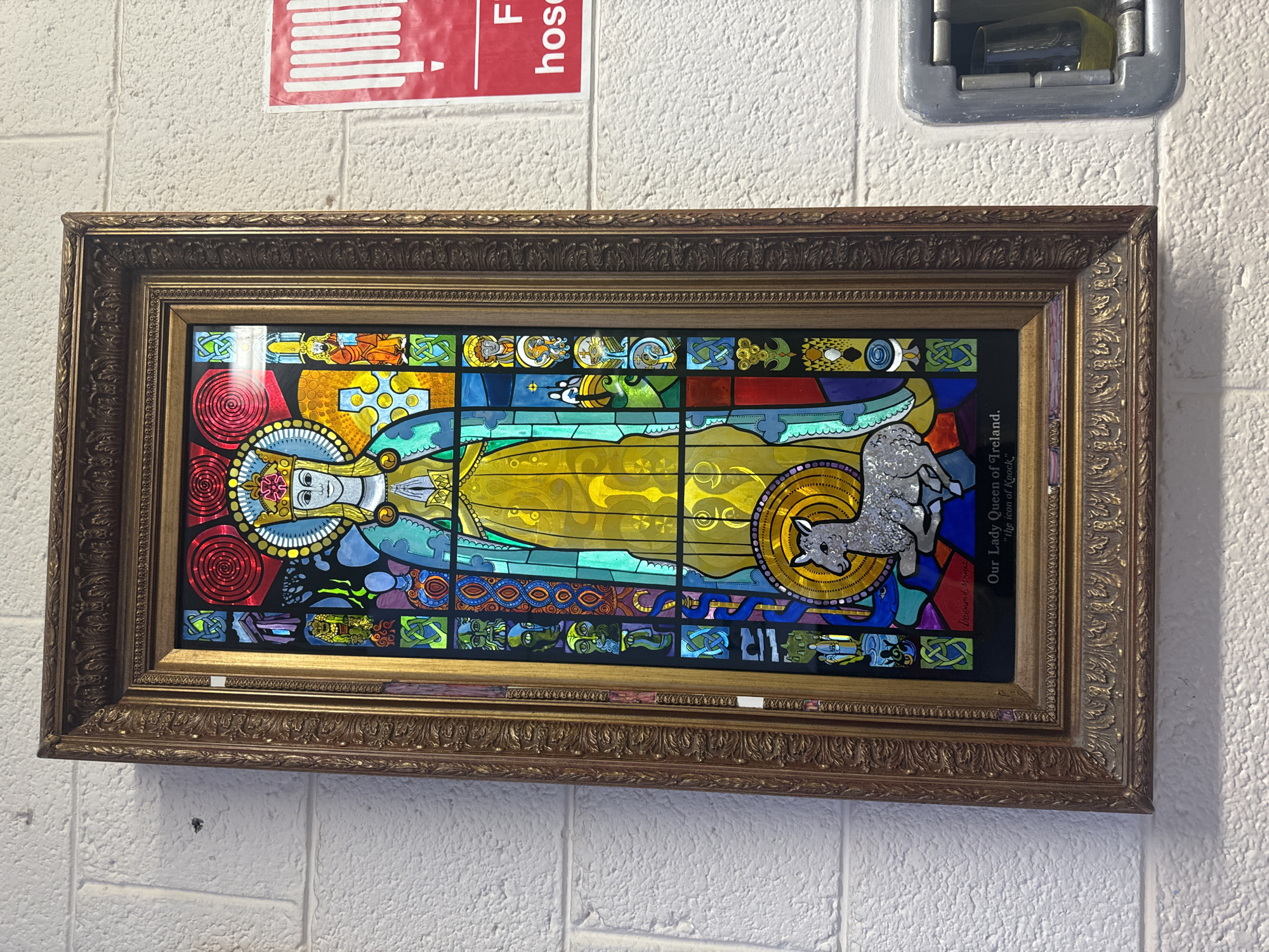 This is a framed stained glass artwork depicting "Our Lady Queen of Ireland, 'the icon of Knock'". The artwork is centered in the frame and shows a depiction of a woman with a crown and halo, wearing a yellow robe and blue mantle. She is shown with her hands clasped in front of her. To her lower left is a lamb with a halo. The stained glass is divided into numerous small panels, many of which feature intricate Celtic knotwork and figures. The frame is ornate and appears to be gold-colored. Below the stained glass, a black plaque reads "Our Lady Queen of Ireland, 'the icon of Knock'". A signature, "Desmond M. Rynes", is visible in small white text on the lower left of the stained glass panel.
The artwork is hanging on a white brick wall. To the right of the artwork, a red sign with white vertical lines is visible, partially obscured by the frame. Below this sign, a metal utility box is mounted on the wall, with a clear glass object inside.
While the prompt requests the location in Waterford, Ireland, this information cannot be determined from the image itself. The image provides no visual cues or textual information that would pinpoint its exact location within Waterford.