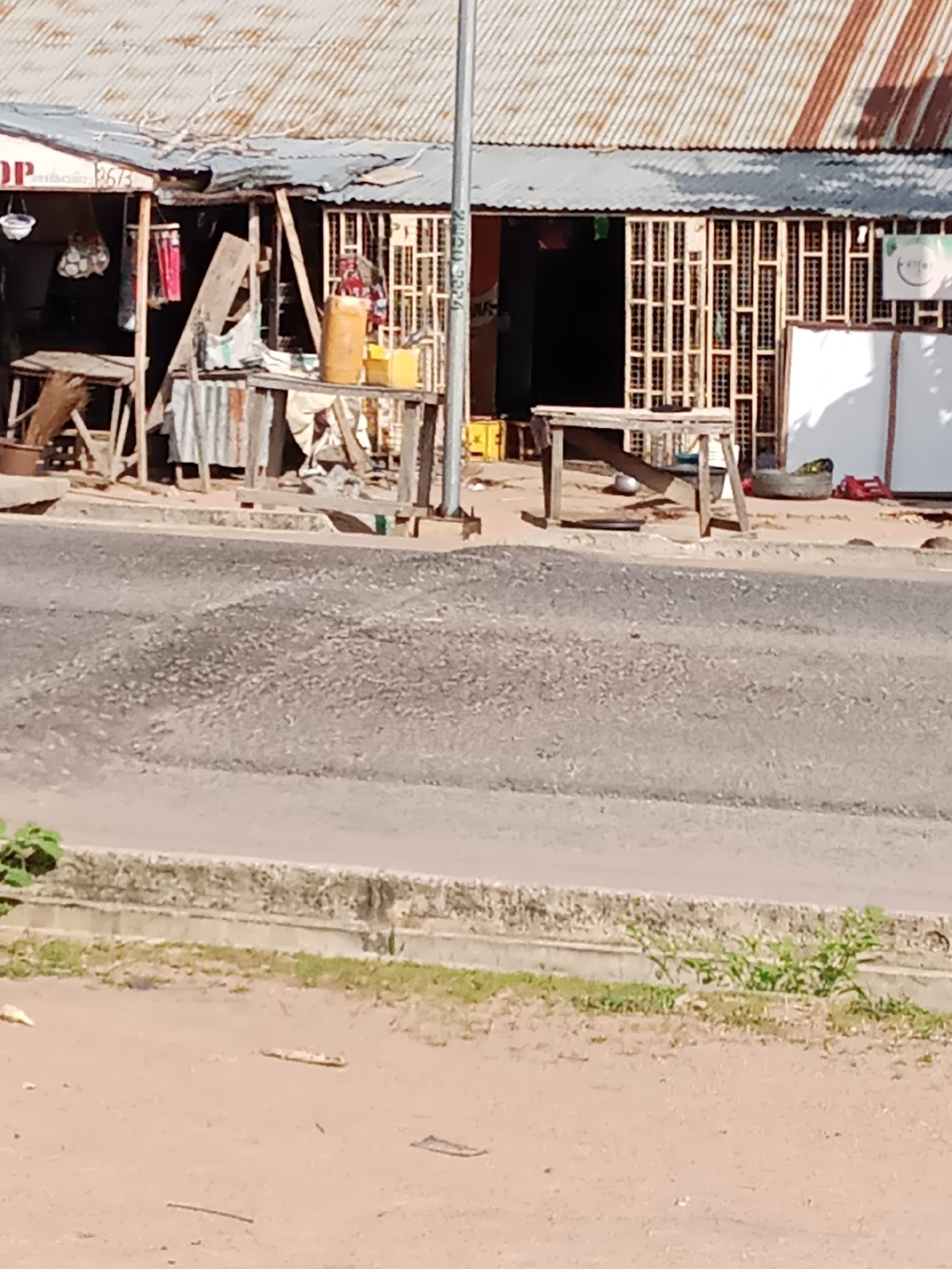 The image depicts a street scene in Jalingo, Nigeria, featuring a row of market stalls with corrugated metal roofing. The stalls are constructed from wood and metal, displaying various items including what appear to be fabrics, containers, and possibly food. A paved road occupies the foreground, separated from the stalls by a concrete curb and a strip of dirt with sparse vegetation. The overall impression is one of an outdoor marketplace or roadside commercial area.