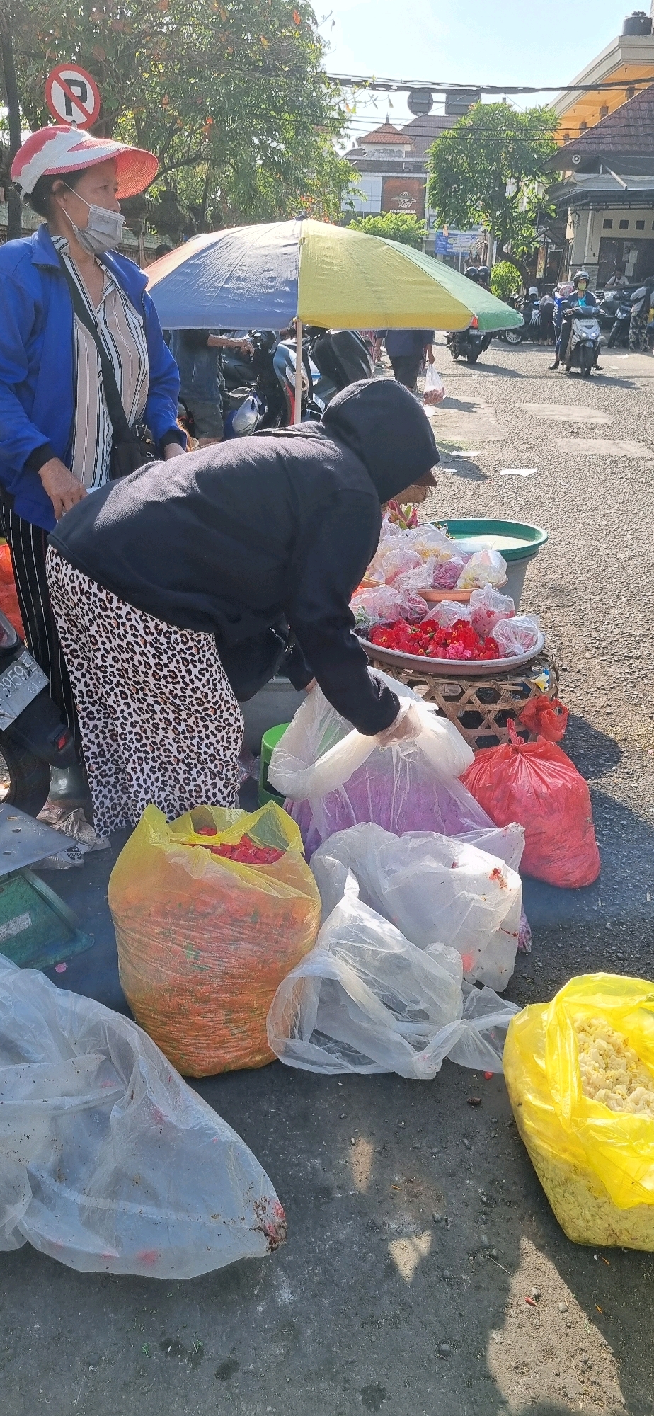 In Denpasar, Indonesia, a street vendor is shown arranging flower offerings at a roadside stall. The vendor, wearing a black hoodie and a leopard-print skirt, is interacting with bags of flowers, likely for religious ceremonies. Behind her, another woman in a blue jacket and a wide-brimmed hat, possibly a customer or fellow vendor, is observing. The scene is set against a backdrop of a street with parked motorcycles and buildings, indicating an urban environment. The presence of colorful flowers in various bags suggests a marketplace or a place where such items are sold. The lighting suggests it is daytime, with bright sunlight casting shadows on the ground.