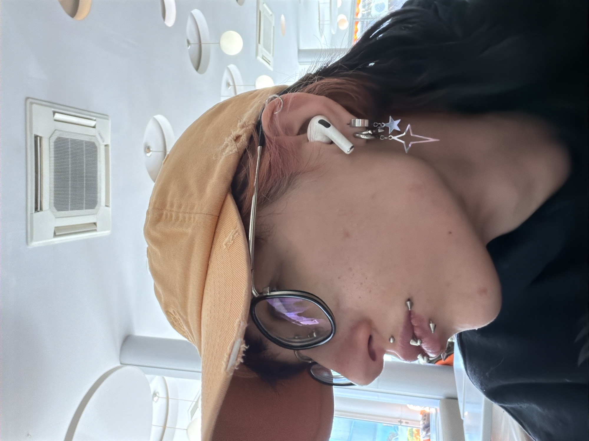 A close-up shot depicts a person wearing a tan baseball cap, eyeglasses, and multiple facial piercings, including piercings on their nose and lip. The person has black hair. An AirPod is visible in their ear, along with other ear jewelry. The background shows a white wall with decorative round lights and a white air conditioning unit. The location of the photo cannot be determined from the provided image.