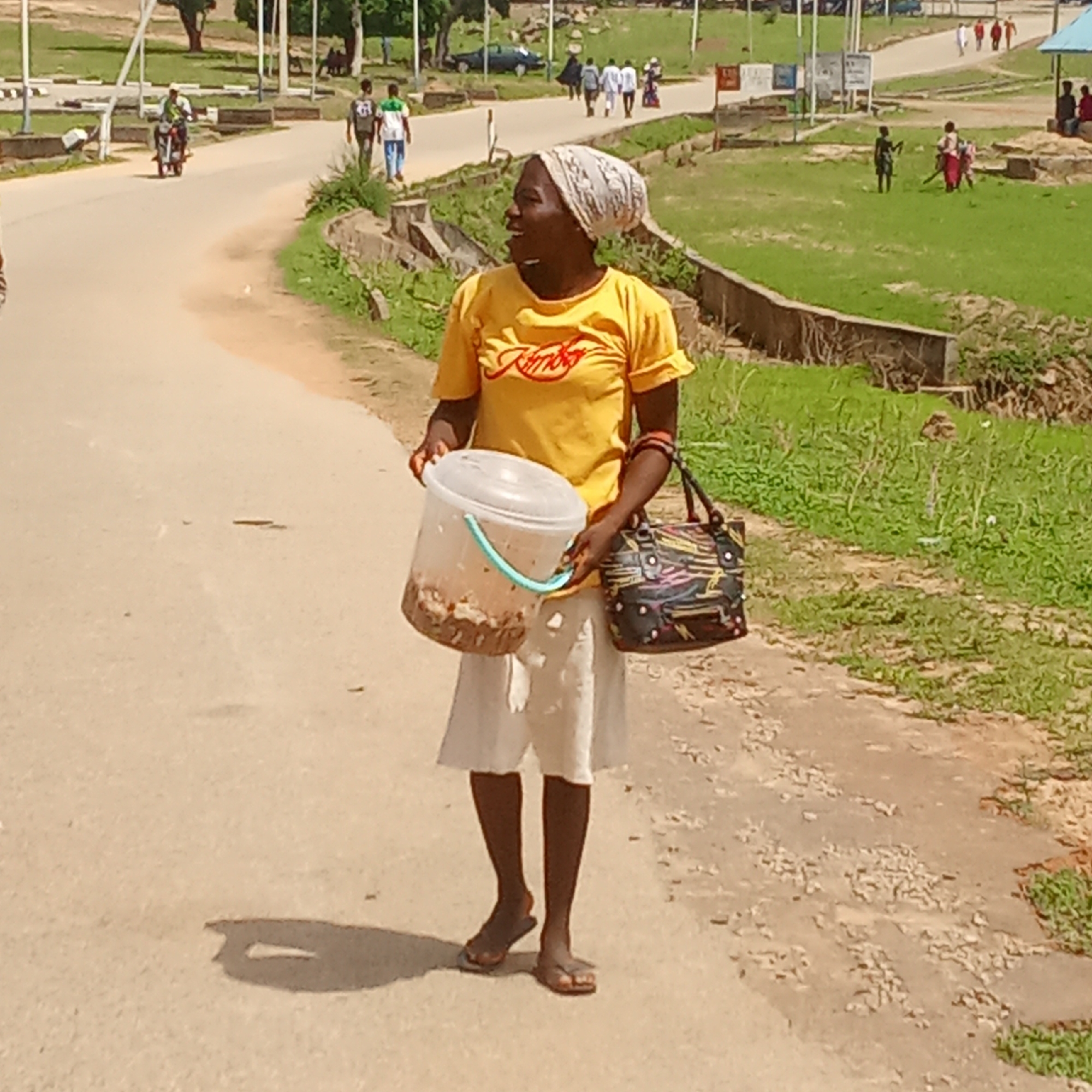 The image captures a woman walking along a paved road in what appears to be a sunny outdoor environment in Akwanga, Nigeria. She is centrally positioned, wearing a bright yellow t-shirt with "Jumbo" printed in red cursive text on the front. Her attire also includes a light-colored headwrap, a knee-length light-colored skirt, and flip-flops.
In her left hand, the woman carries a transparent plastic bucket that contains dark, granular items, possibly food or snacks, suggesting she might be a street vendor. In her right hand, she holds a multi-colored handbag with a vibrant, abstract pattern. Her face is turned to her left, and she has a broad smile, indicating a cheerful or contented mood.
The setting includes a curved asphalt road with visible shadows, suggesting bright sunlight, likely indicating midday or early afternoon. To the left of the road, a person on a motorcycle and several individuals walking are visible in the distance. To the right, beyond the road's edge, there is a grassy area with some sparse vegetation and concrete structures. Further in the background, a few children are seen on a larger grassy expanse, with more adults walking along another section of the road. The overall scene depicts everyday activity in a lively, communal space.