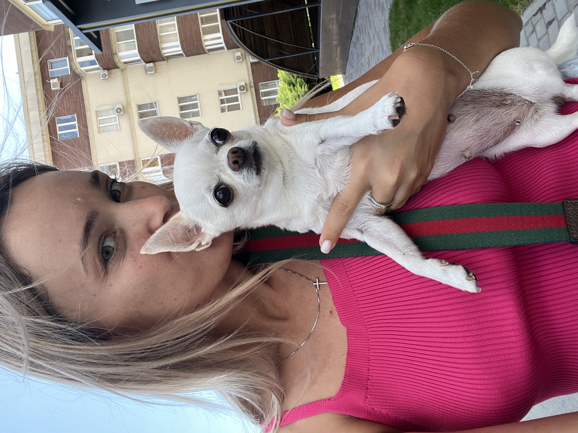 In Kryzhanivka, Ukraine, a woman with blonde hair, wearing a bright pink ribbed tank top and a silver necklace with a cross pendant, is taking a selfie outdoors. She is holding a small white chihuahua dog, which is looking directly at the camera with large dark eyes. The dog has its ears perked up. Behind them, a multi-story residential building with beige and brown facades and visible air conditioning units is visible. The background also includes a paved walkway, green grass, and a covered seating area. A green and red striped strap, possibly part of a bag, is draped across the woman's shoulder and chest.